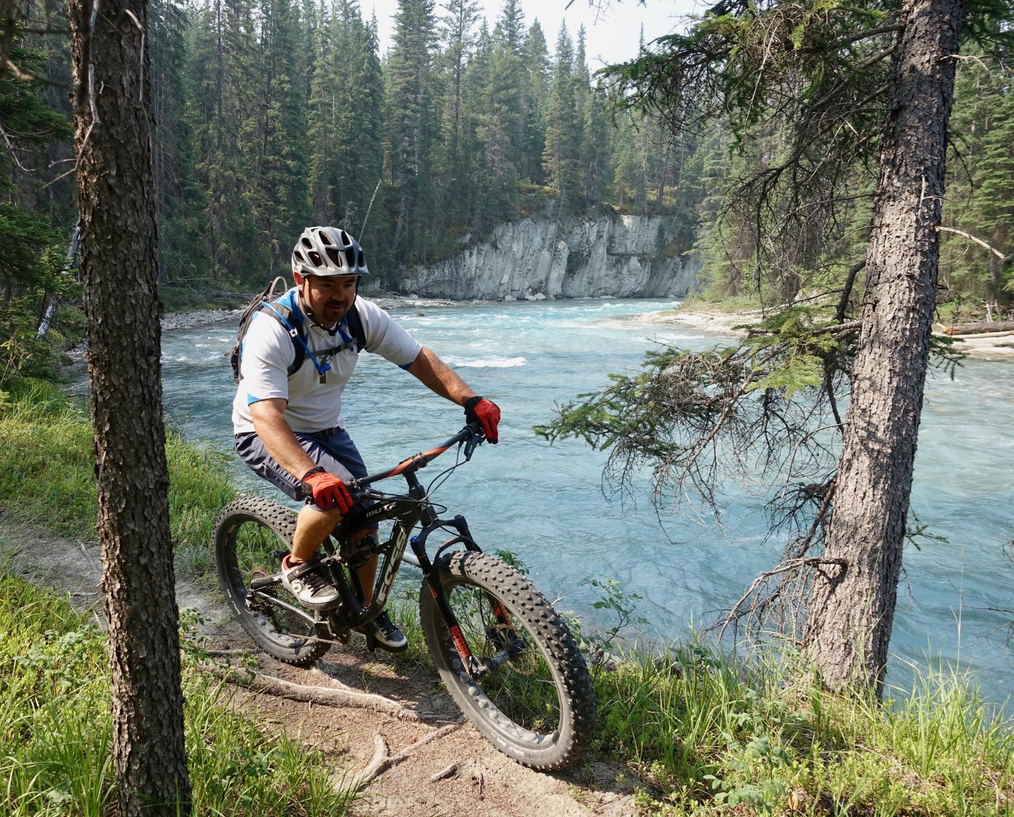 A mountain biker navigating a narrow dirt trail along a river, surrounded by tall green trees and rocky cliffs. The rider is wearing a helmet and gloves, appearing focused as he maneuvers his bike near the water's edge. Nipika Mountain Resort mountain bike trail.