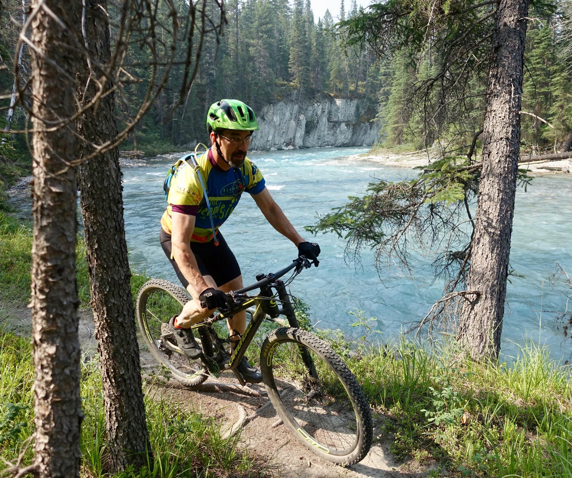 A mountain biker navigates a dirt path beside a turquoise river, surrounded by tall pine trees. The cyclist is wearing a colorful jersey and a bright green helmet. Sunlight filters through the trees, creating a picturesque outdoor scene. Nipika Mountain Resort mountain bike trail.