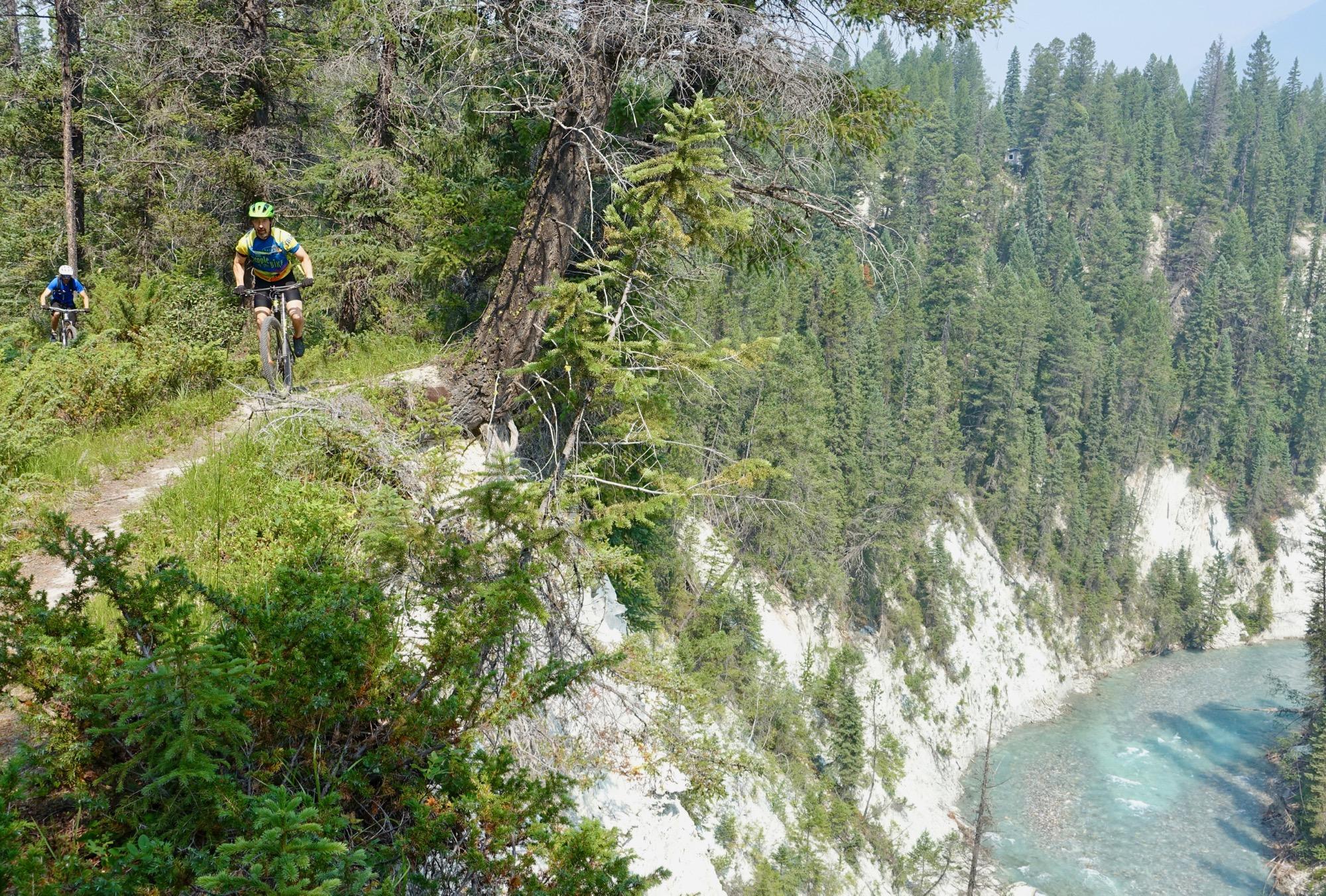 A person riding a mountain bike on a narrow dirt trail surrounded by lush greenery, with a steep cliff and a river visible below. Another cyclist can be seen in the background. The scene captures a sunny day in a mountainous area, emphasizing the adventurous spirit of outdoor biking. Nipika Mountain Resort mountain bike trail.