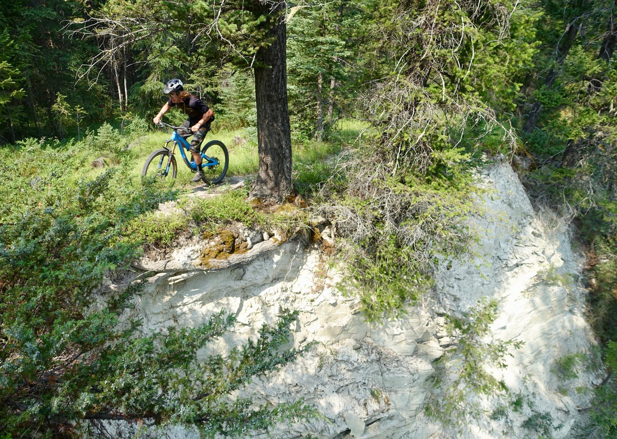 A mountain biker navigating a narrow trail next to a steep, rocky cliff surrounded by lush greenery and trees in a forested area. Nipika Mountain Resort mountain bike trail.