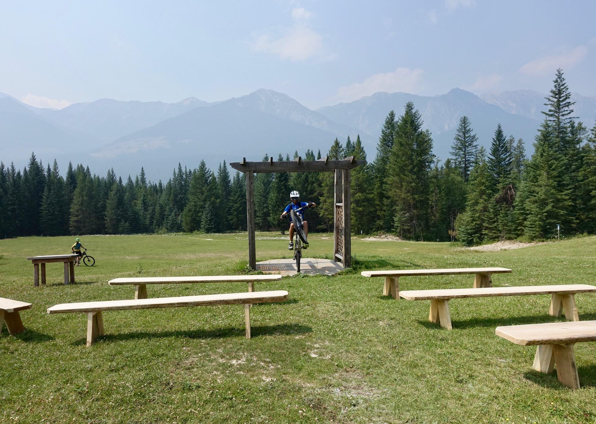 A mountain biker performing a trick while jumping through a wooden arch structure in a grassy open area, with benches arranged in the foreground and a backdrop of mountains and trees. The sky is clear and slightly hazy. Nipika Mountain Resort mountain bike trail.
