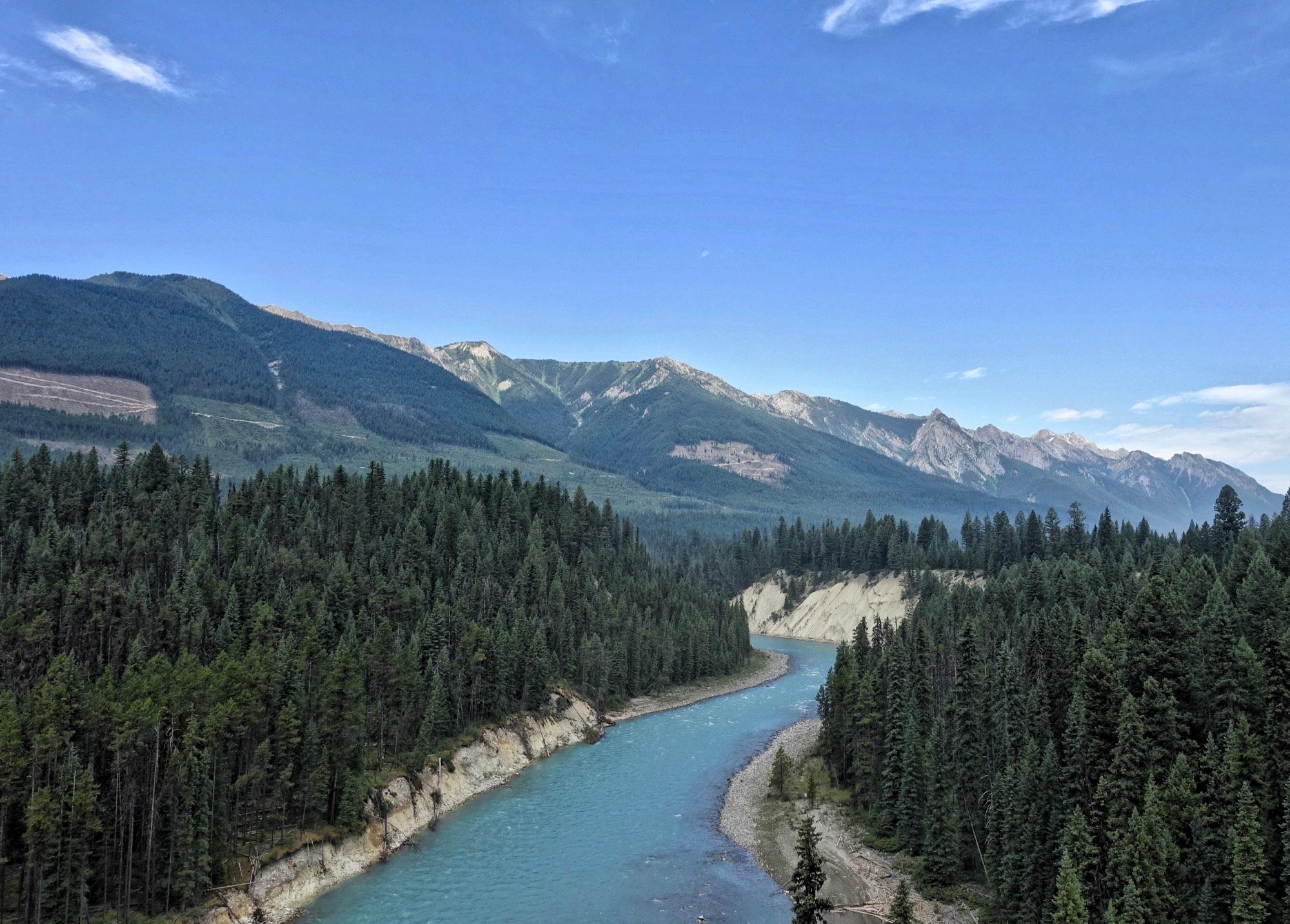 A scenic view of a winding river surrounded by lush green forests and towering mountains under a bright blue sky. The river reflects varying shades of turquoise as it flows through the landscape, with steep cliffs and rocky banks visible in certain areas.  Nipika Mountain Resort mountain bike trail.