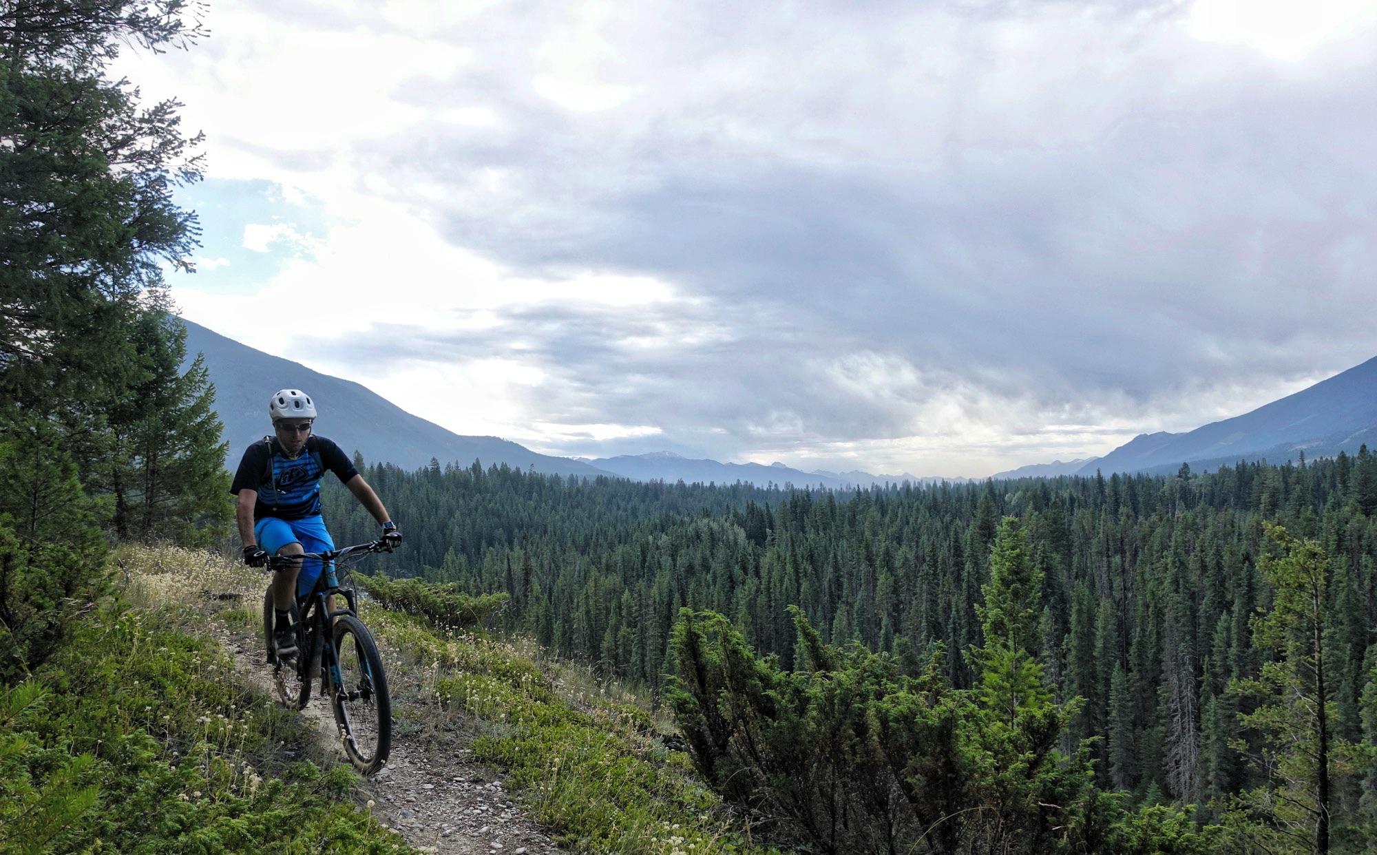 A mountain biker riding along a dirt trail bordered by lush greenery, with a panoramic view of dense forests and mountains in the background under a cloudy sky. Nipika Mountain Resort mountain bike trail.