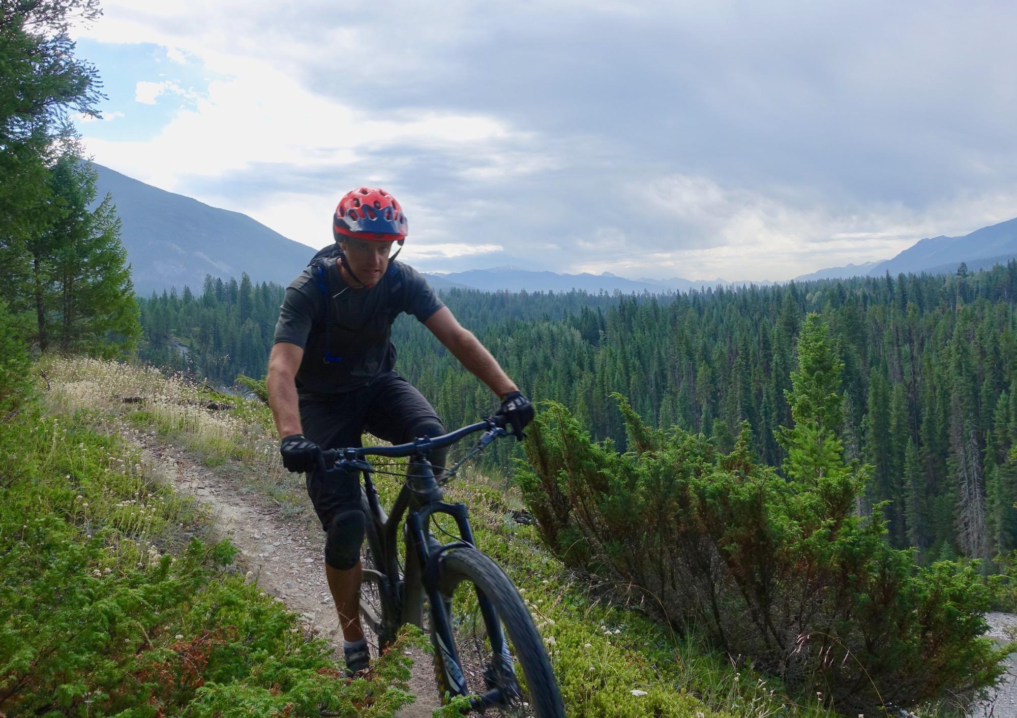A mountain biker navigates a narrow trail along a scenic hillside, surrounded by lush greenery and mountains in the background under a cloudy sky. The rider is wearing a red helmet and black attire, focused on the path ahead. Nipika Mountain Resort mountain bike trail.