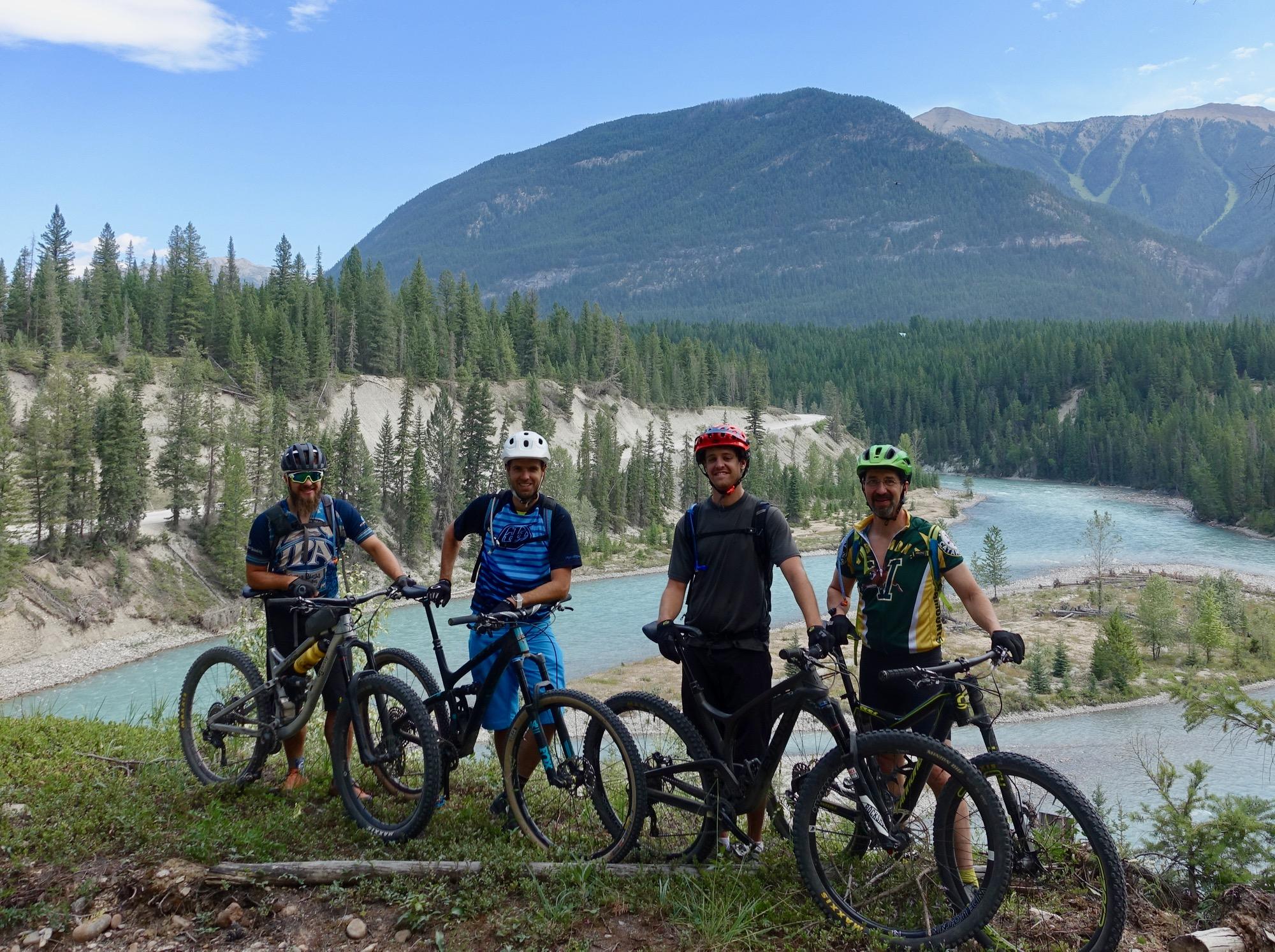 Four mountain bikers pose together on a hillside overlooking a scenic river surrounded by pine trees and mountains. They are wearing helmets and riding gear, standing beside their bikes. The sky is clear with a few clouds. Nipika Mountain Resort mountain bike trail.