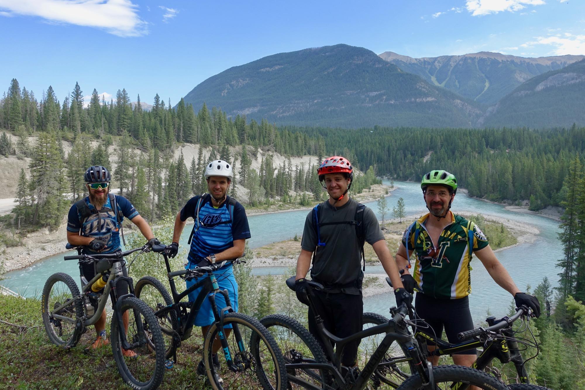Four mountain bikers pose for a photo on a hillside, with a river and dense forest in the background. They are wearing helmets and cycling gear, standing beside their bikes. The scenery features mountains in the distance under a clear blue sky. Nipika Mountain Resort mountain bike trail.