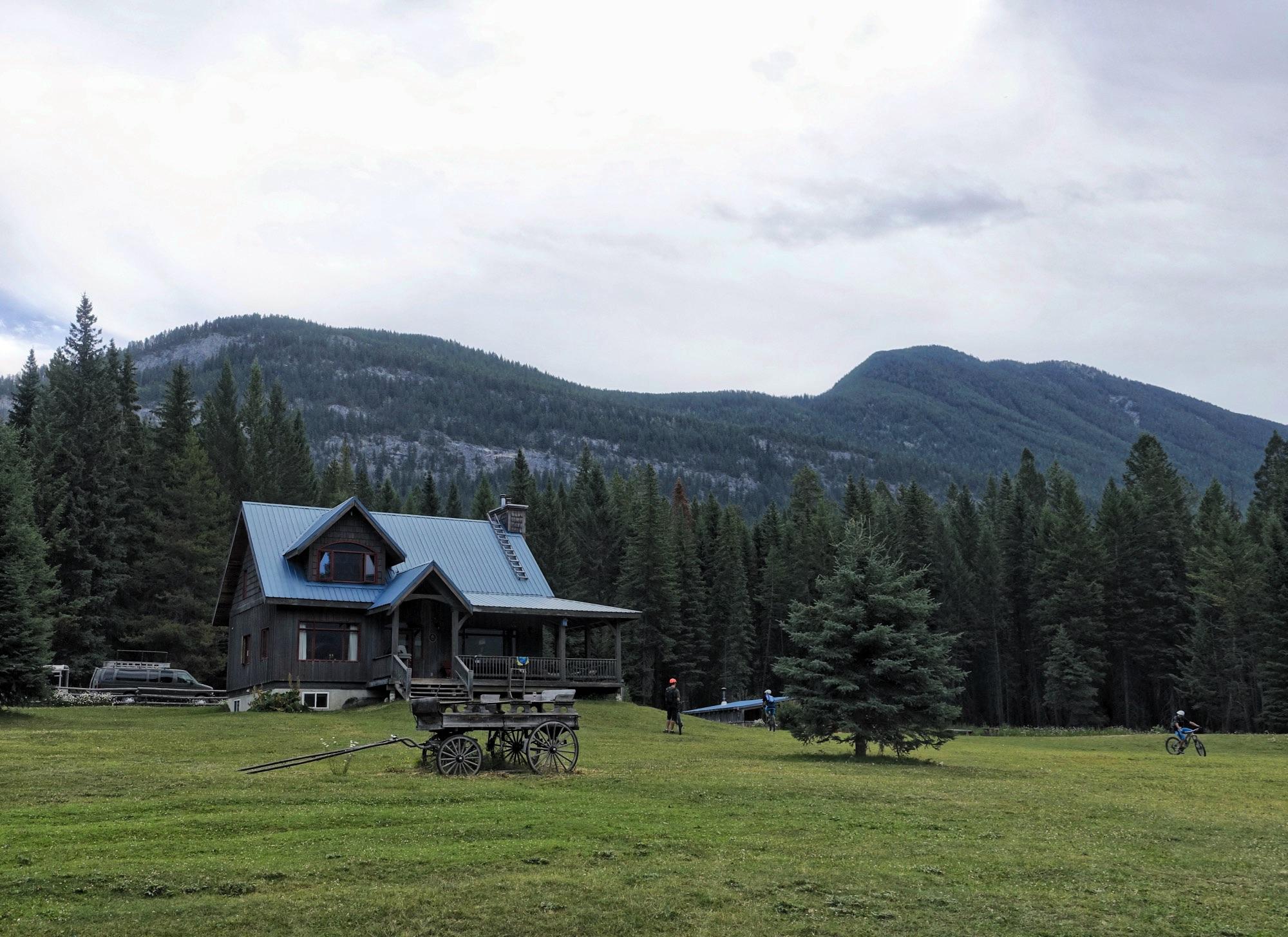 A scenic view of a rustic cabin with a blue metal roof, surrounded by lush green grass and tall trees. In the foreground, an old wooden cart is parked on the lawn. Two people are visible in the area, one near a tree and another riding a bicycle in the background. Majestic mountains are seen in the distance under a cloudy sky. Nipika Mountain Resort mountain bike trail.