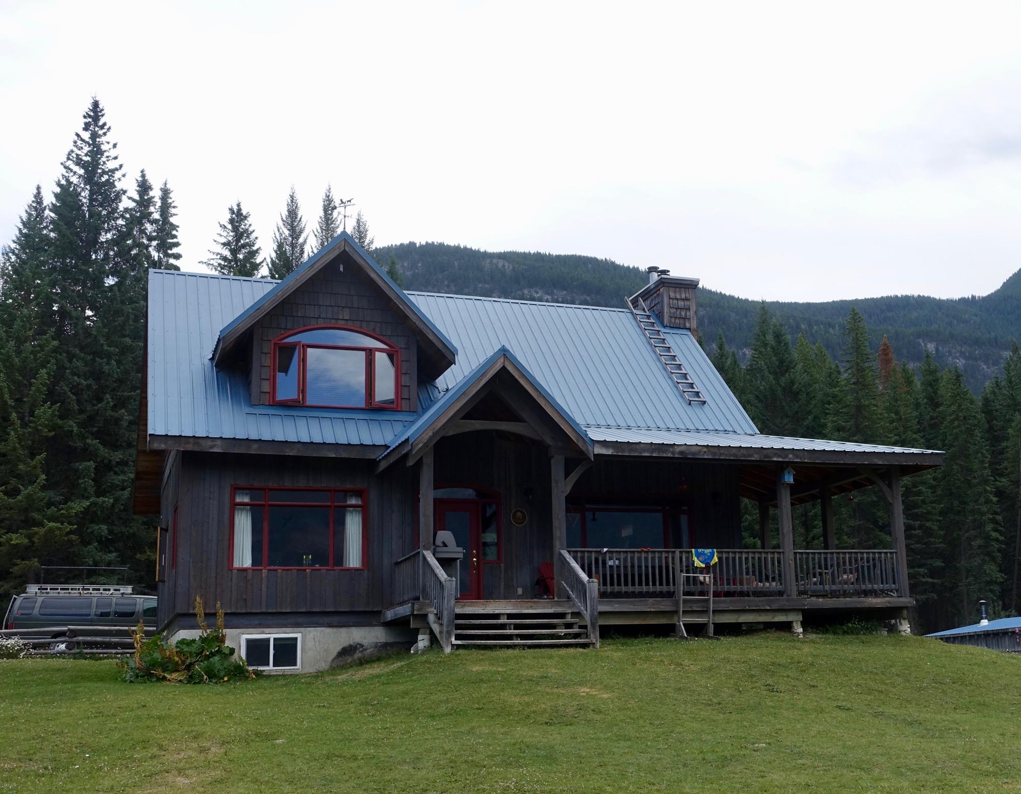 A rustic wooden house with a blue metal roof, featuring red-trimmed windows and a welcoming front porch. Surrounded by lush green grass and tall evergreen trees, the house sits in a mountainous landscape under a cloudy sky. A vehicle can be seen parked next to the house. Nipika Mountain Resort mountain bike trail.