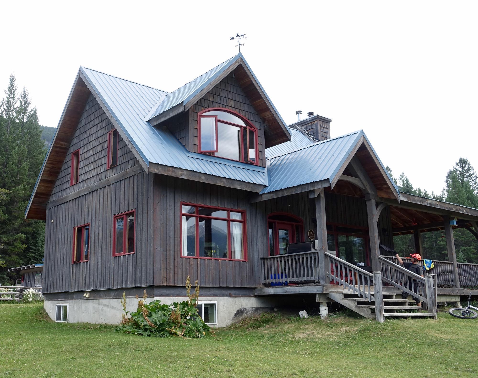 A large wooden house with a blue metal roof and red-framed windows, situated on a grassy area. It features a spacious porch with wooden steps and a railing, surrounded by trees in the background. Nipika Mountain Resort mountain bike trail.