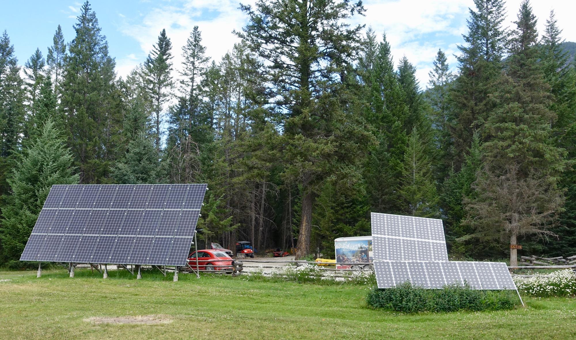 A grassy area featuring two solar panel arrays positioned among tall trees. In the background, there are parked vehicles and a sign partially visible. The scene is set under a partly cloudy sky, highlighting the natural surroundings. Nipika Mountain Resort mountain bike trail.