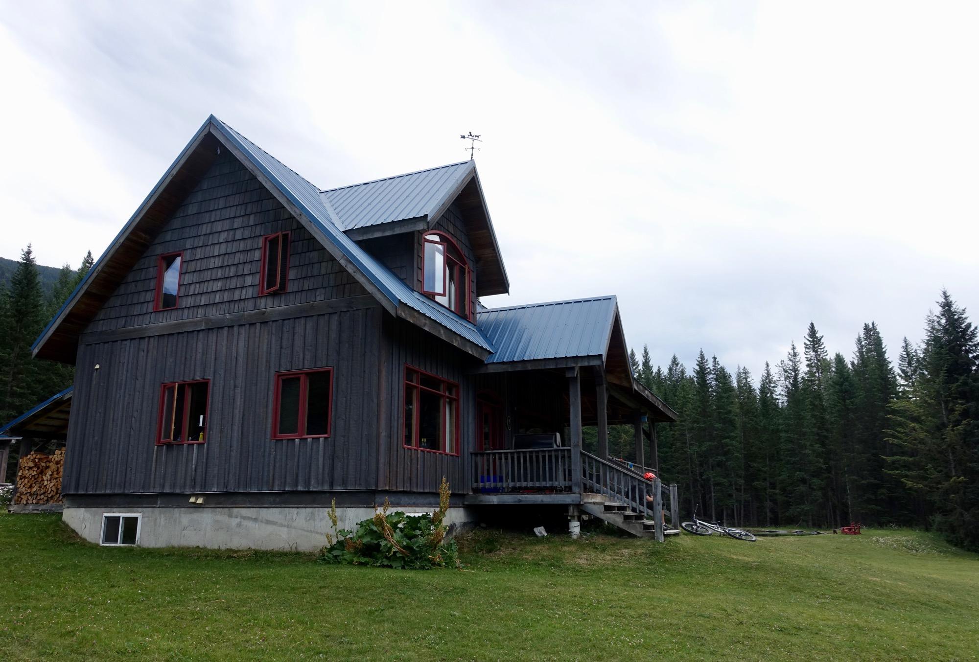 A rustic wooden house with a blue metal roof, featuring red windows and a front porch, surrounded by lush green grass and tall evergreen trees under a cloudy sky. Nipika Mountain Resort mountain bike trail.