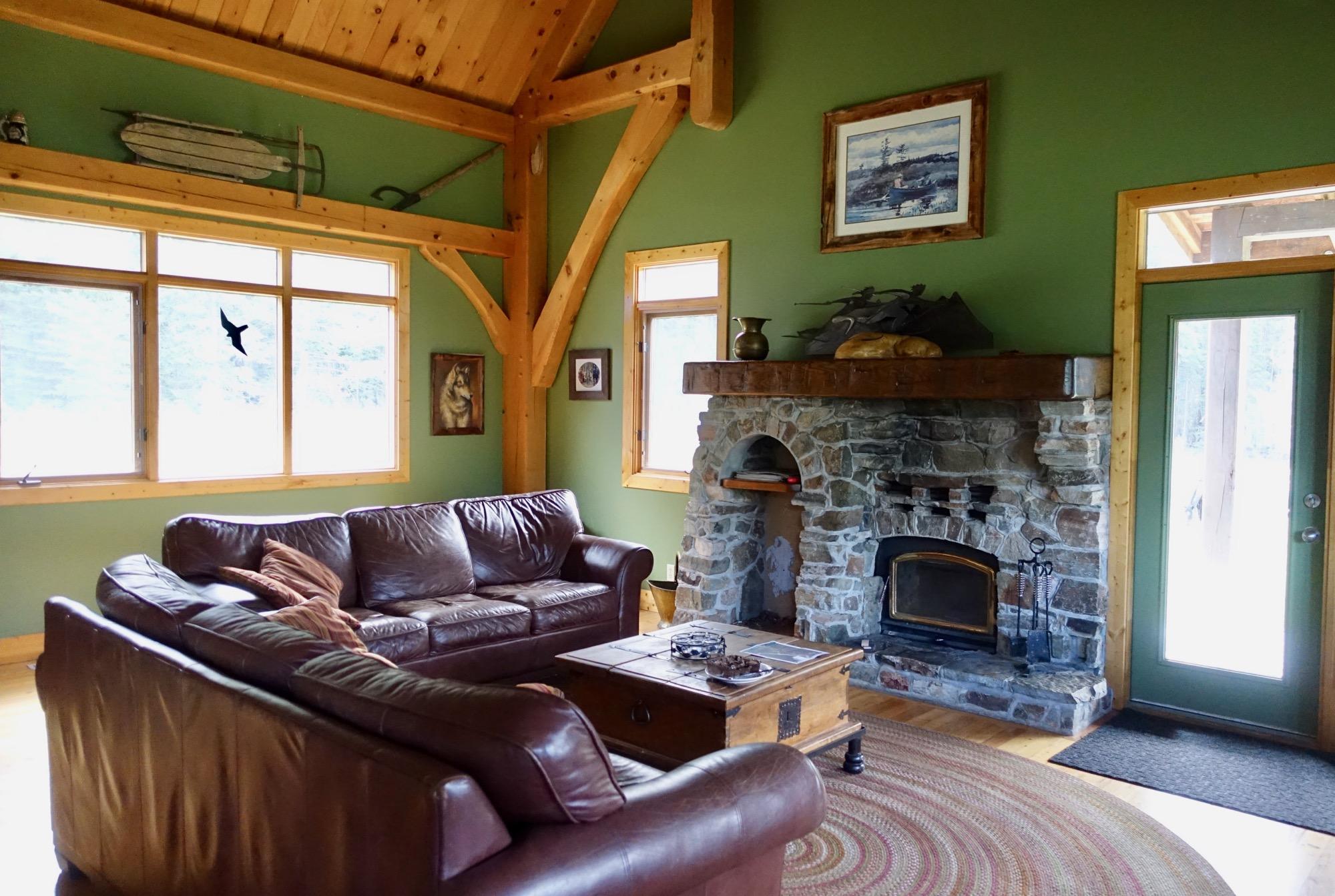 Cozy living room with a stone fireplace, leather sofas, and wooden beams. Large windows provide natural light, and rustic decor, including artwork and sleds, adorns the walls. A round braided rug and a wooden coffee table complete the inviting atmosphere. Nipika Mountain Resort mountain bike trail.