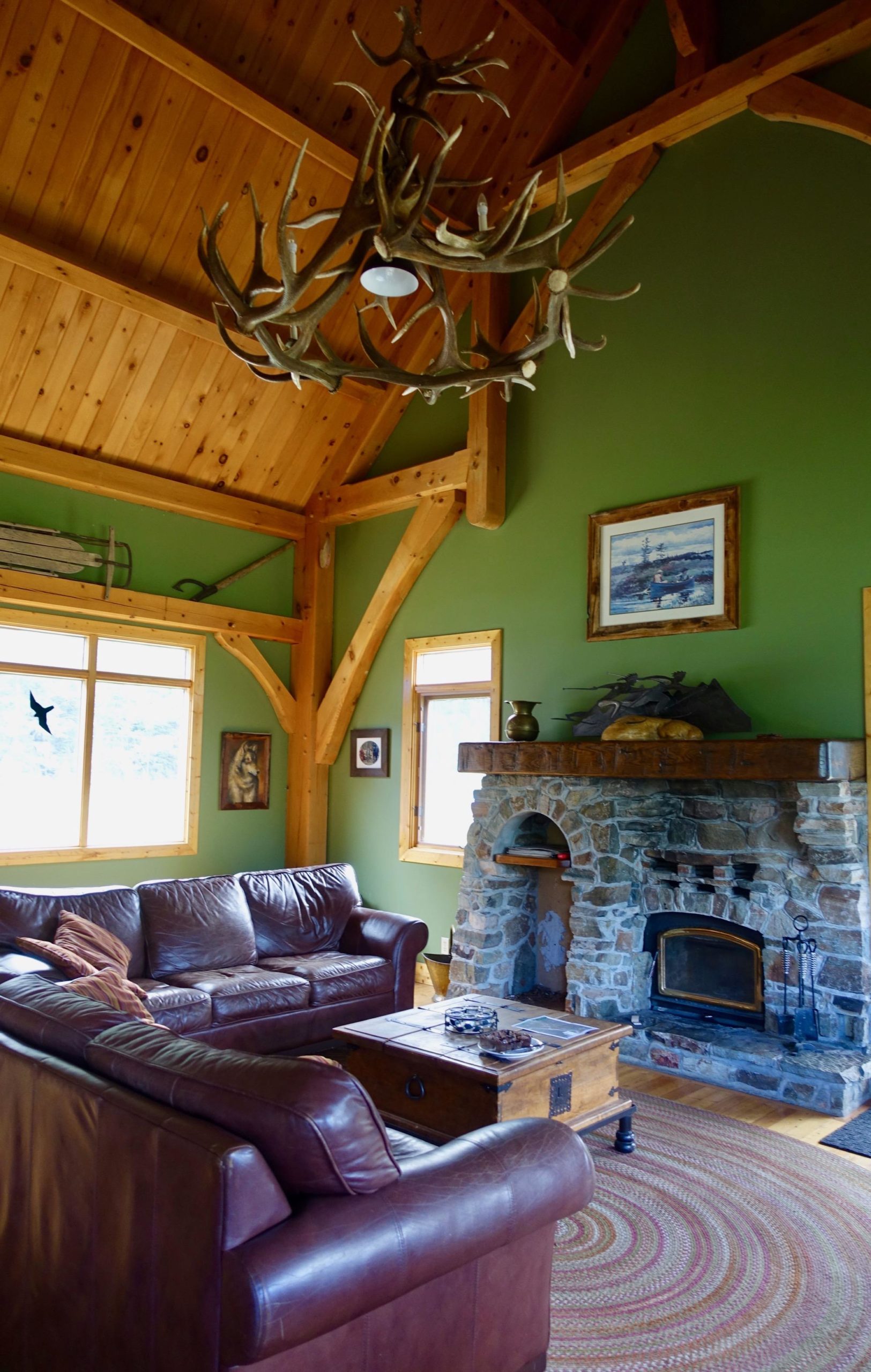 A cozy living room featuring a leather sofa set, a wooden coffee table, and a stone fireplace. The walls are painted a calming green, and there are large windows allowing natural light to fill the space. The ceiling is made of wooden beams, and a unique antler chandelier hangs overhead. Various artworks and decorative items adorn the walls, contributing to a warm, rustic atmosphere. Nipika Mountain Resort mountain bike trail.