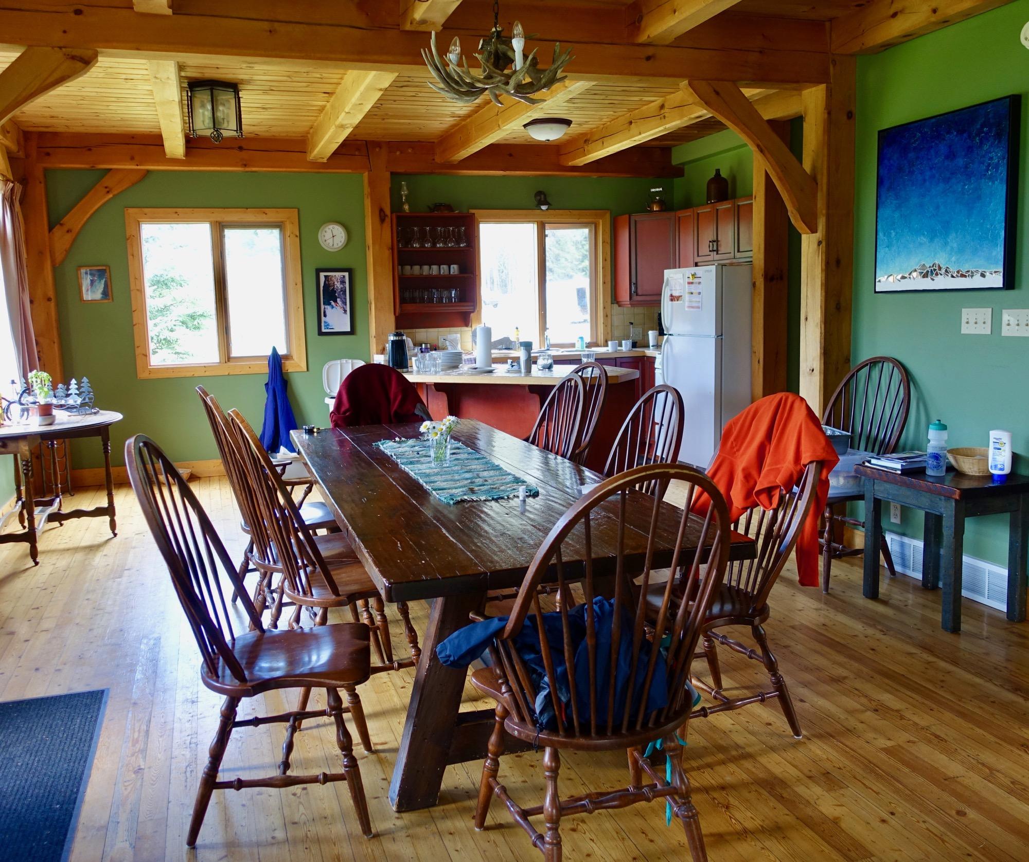 Cozy, rustic dining area featuring a large wooden table surrounded by wooden chairs. The walls are painted in a soothing green, complemented by wooden beams and a natural wood floor. Sunlight filters through large windows, illuminating a kitchen area with red cabinets and essential appliances. A few personal items, including clothing and a water bottle, are casually placed around the space, adding a lived-in feel. Nipika Mountain Resort mountain bike trail.