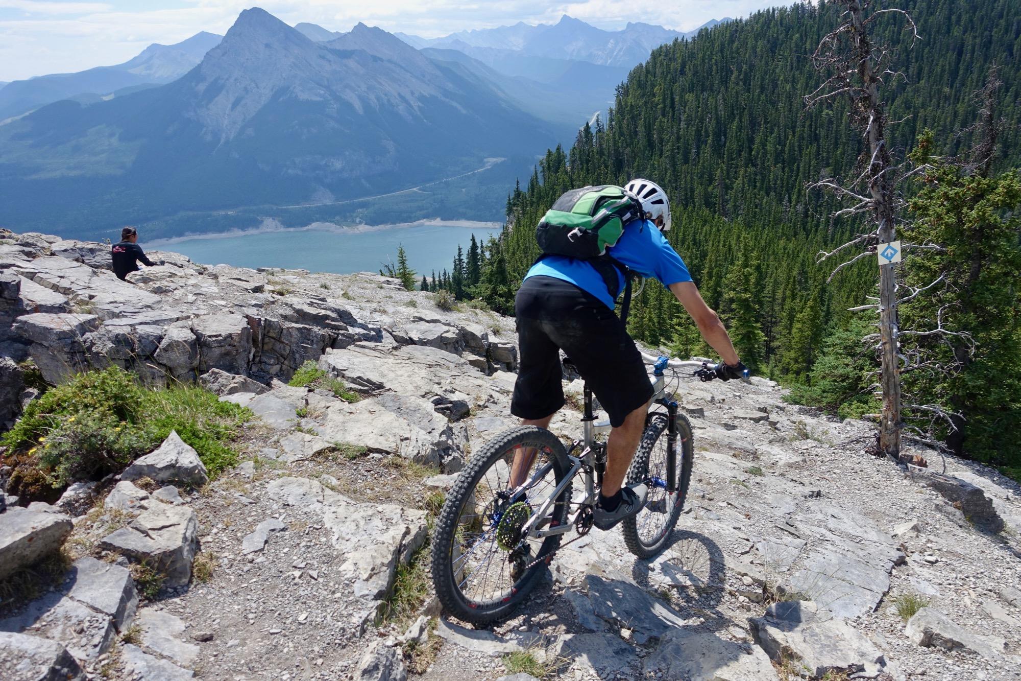 A mountain biker riding down a rocky trail with a scenic view of mountains and a lake in the background. The biker is wearing a blue shirt and a helmet, and there are lush green trees surrounding the area. A person is sitting on a rock in the distance, enjoying the view. Jewell Pass Trail mountain bike trail.