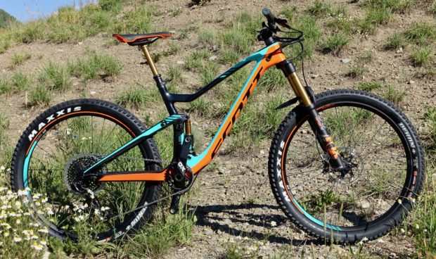 A brightly colored mountain bike with a teal and orange frame, featuring wide tires with "Maxxis" branding, is positioned on a rocky surface surrounded by green grass and wildflowers. The bike showcases a modern design with a prominent front suspension fork and a sleek saddle.