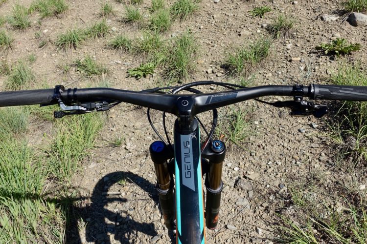 Alt text: A top-down view of a mountain bike handlebar, featuring textured grips and gear shift levers, with grass and rocky ground visible in the background.