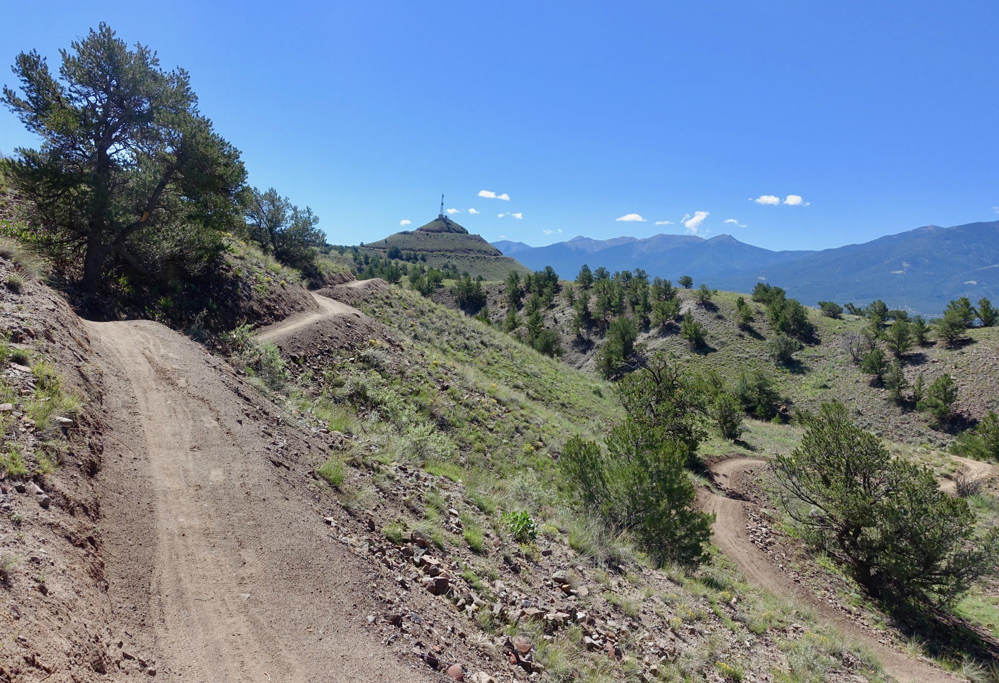 A winding dirt trail leads through a hilly landscape, with green shrubs and trees flanking the path. In the distance, a peak with a communication tower is visible, set against a clear blue sky with a few scattered clouds. Mountains stretch across the background, creating a picturesque natural scene. Rusty Lung mountain bike trail.