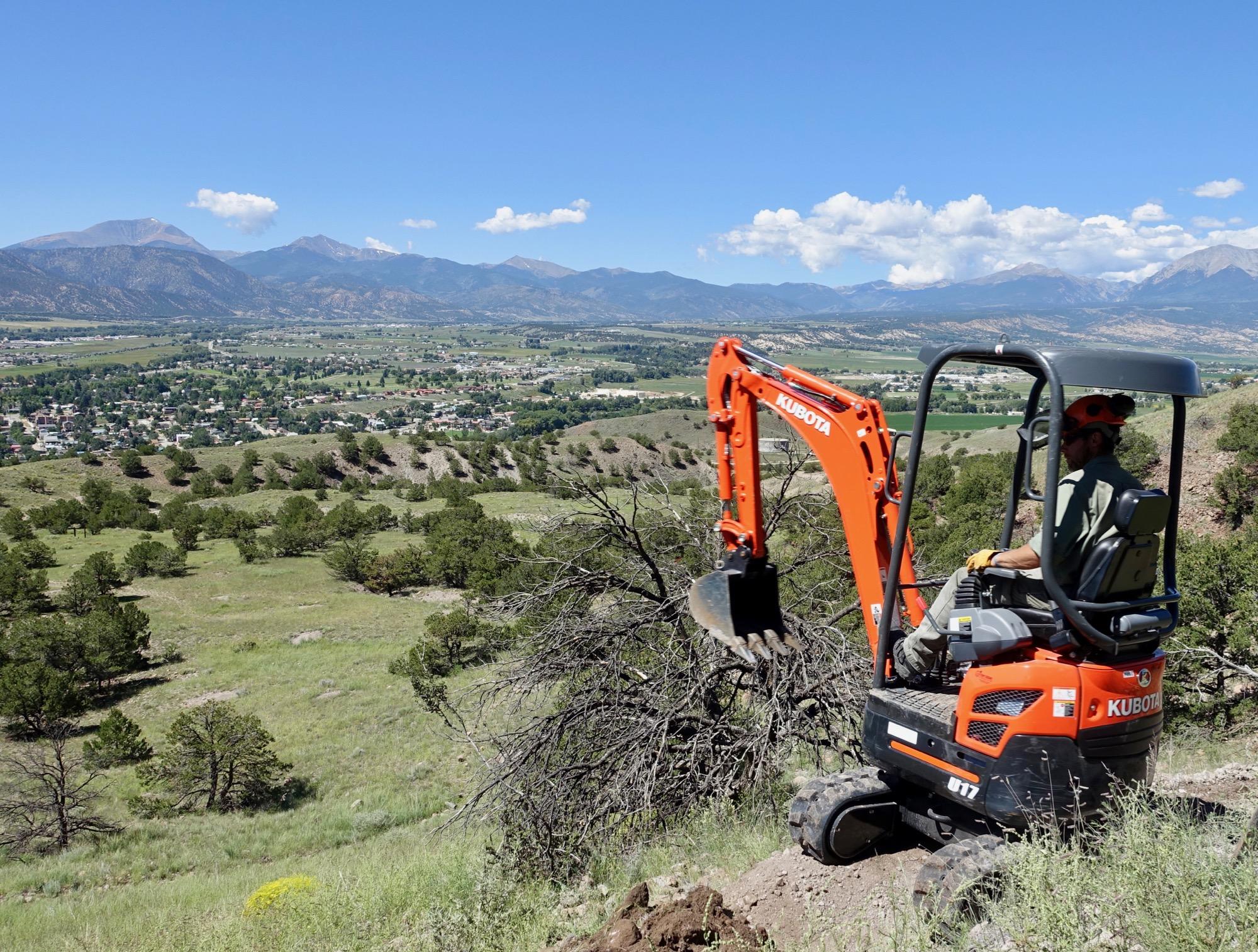 A person operating a small excavator on a hillside, overlooking a valley with a town and mountains in the background. The scene features a clear blue sky and lush green vegetation. The excavator is orange, and the operator is wearing a hard hat and gloves. Arkansas Hills mountain bike trail.