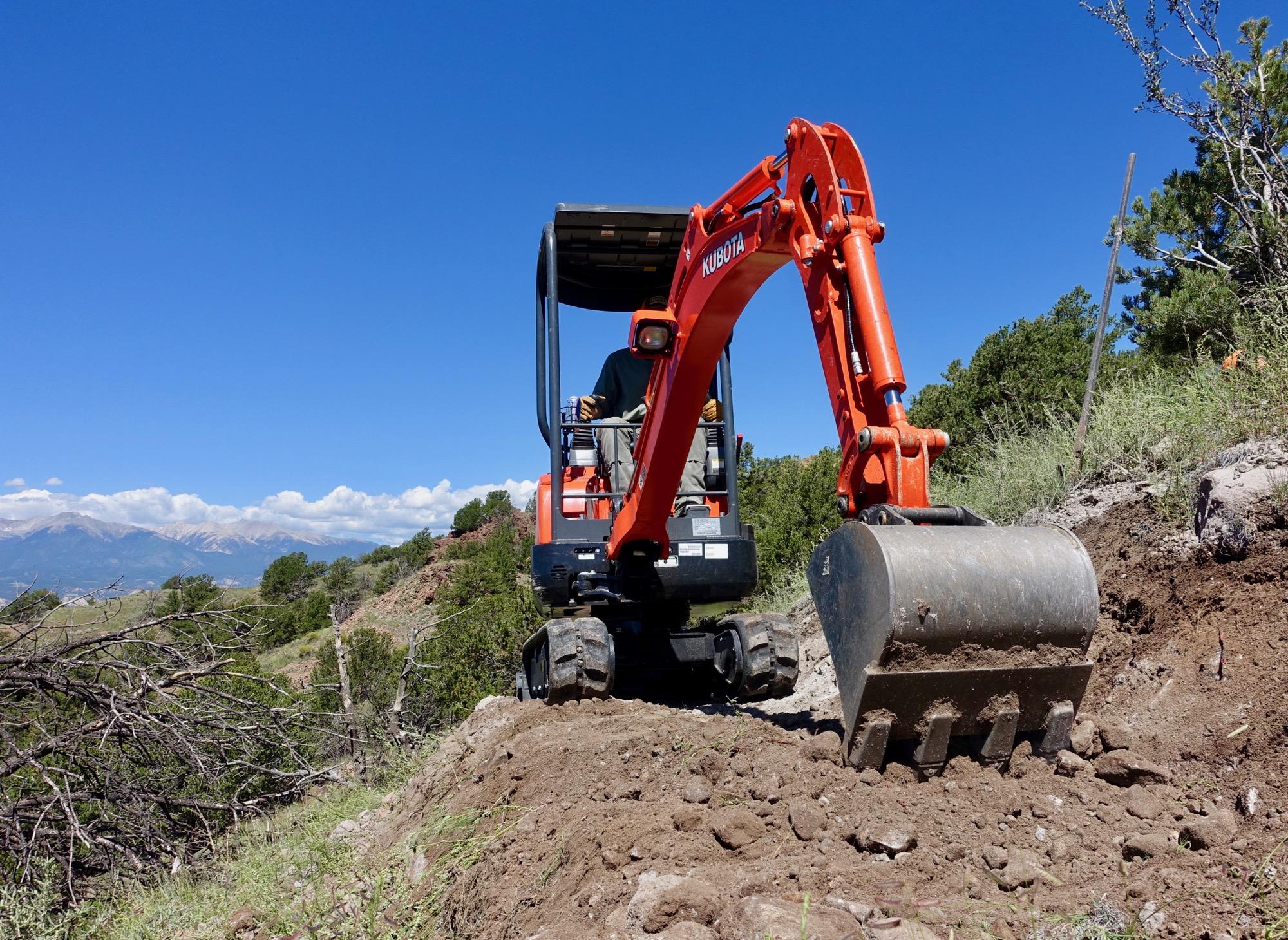 A small orange excavator working on a construction site, with a clear blue sky and distant mountains in the background. The excavator is positioned on a dirt hillside surrounded by greenery. Arkansas Hills mountain bike trail.