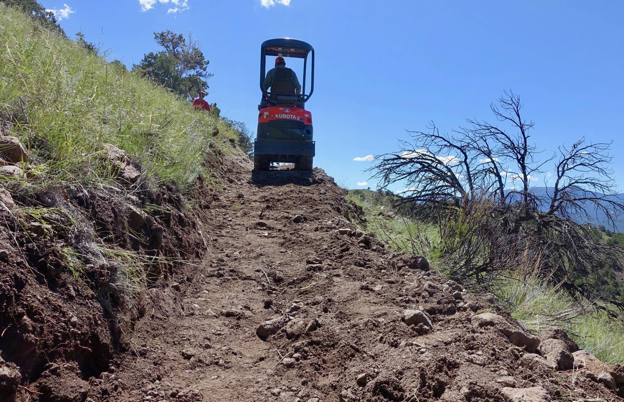 A small excavator or bulldozer working on a narrow dirt path through hilly terrain, surrounded by grass and trees under a bright blue sky. Arkansas Hills mountain bike trail.