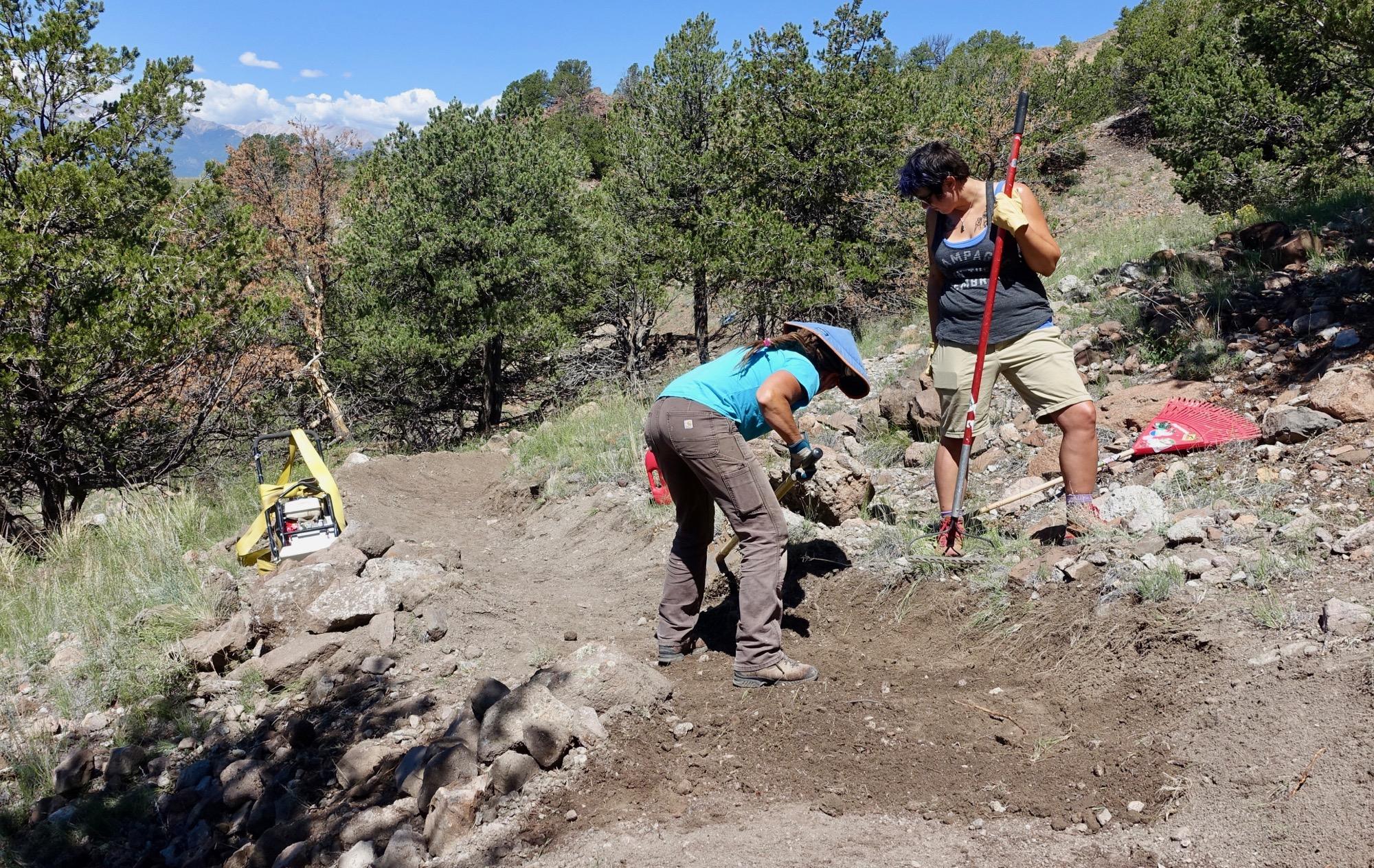 Two individuals are working on a trail-building project in a forested area. One person, wearing a blue shirt and a wide-brimmed hat, is using a tool to dig into the ground, while the other, dressed in a tank top and shorts, stands nearby with a rake. In the background, there are trees and rocky terrain, and construction equipment is visible on the side. The scene is set under a clear blue sky. Arkansas Hills mountain bike trail.