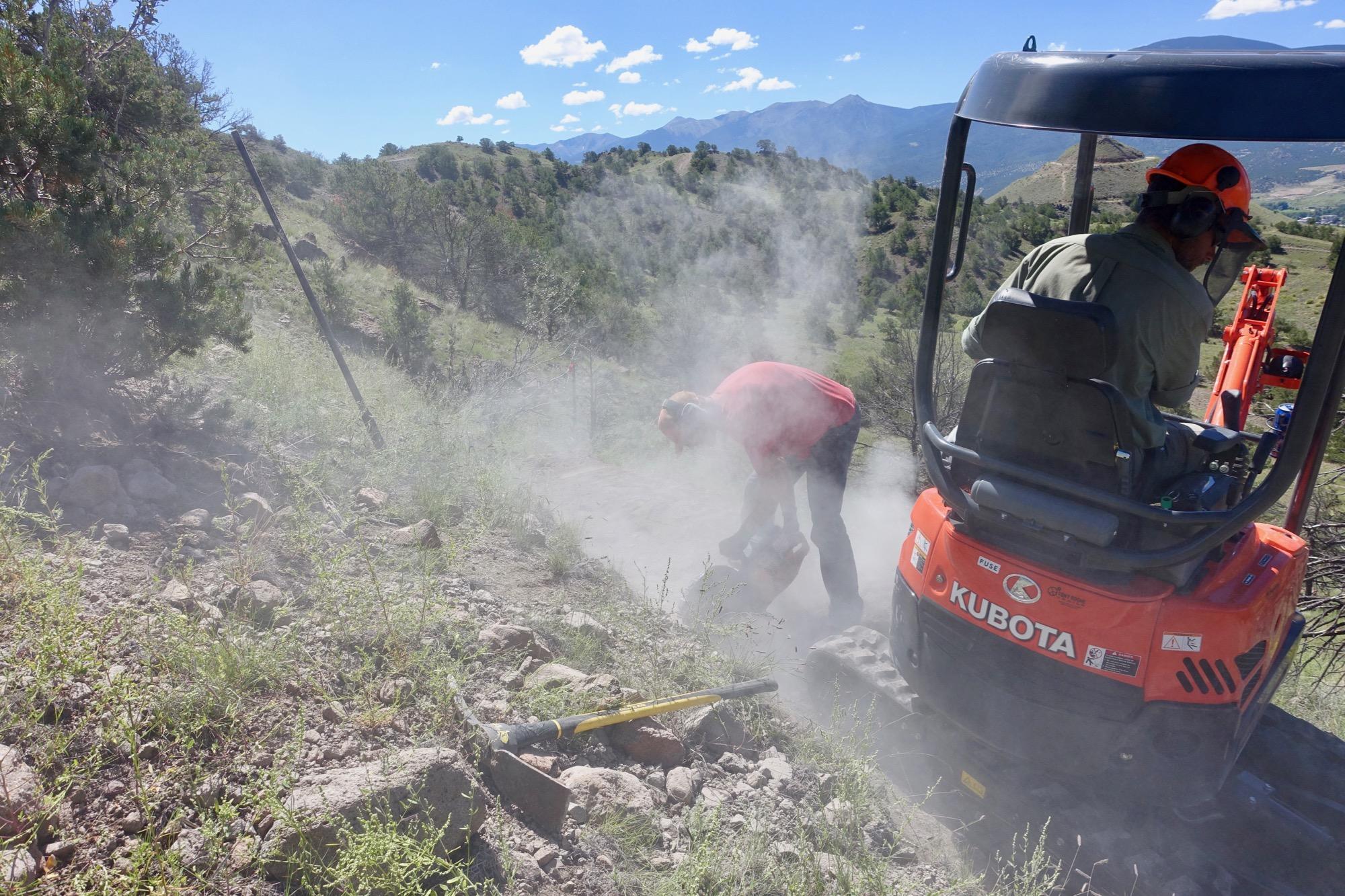 Two workers are engaged in a landscaping project on a hillside. One worker, wearing a red shirt and ear protection, is using a tool near the ground, creating dust. The other worker is seated in a small orange Kubota machine, operating it from the right side of the image. The background features a green, hilly landscape with distant mountains and a blue sky dotted with clouds. Arkansas Hills mountain bike trail.