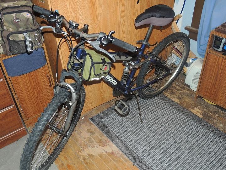 Mongoose Mongoose Ledge 2.1: A mountain bike leaning against a wooden cabinet, featuring a green gear bag attached to the front frame. The bike is positioned on a textured rug, with wooden flooring visible around it. In the background, a small piece of furniture and a window with blue curtains can be seen.
