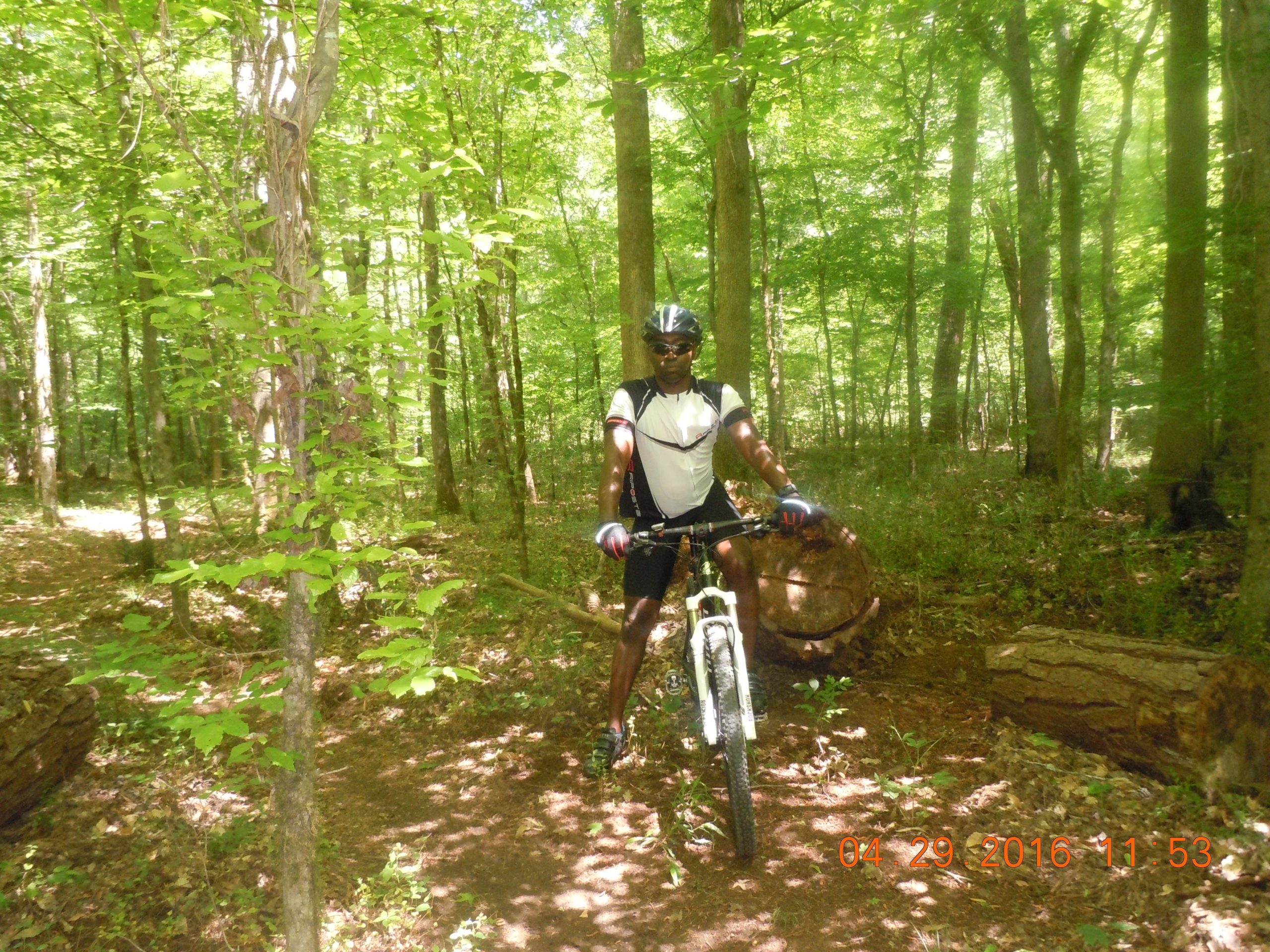 A person wearing a helmet and cycling gear stands next to a mountain bike on a dirt trail surrounded by lush green trees in a forested area. Sunlight filters through the leaves, creating a serene outdoor atmosphere. Dauset Trails Nature Center mountain bike trail.