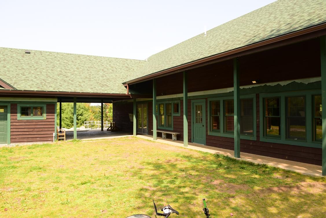 A view of a rustic building with dark wood siding and green accents, featuring a sloped green roof. The image captures an outdoor area with a grassy lawn, several windows, and a porch area, surrounded by trees in the background under a clear sky. Dewey Mountain mountain bike trail.