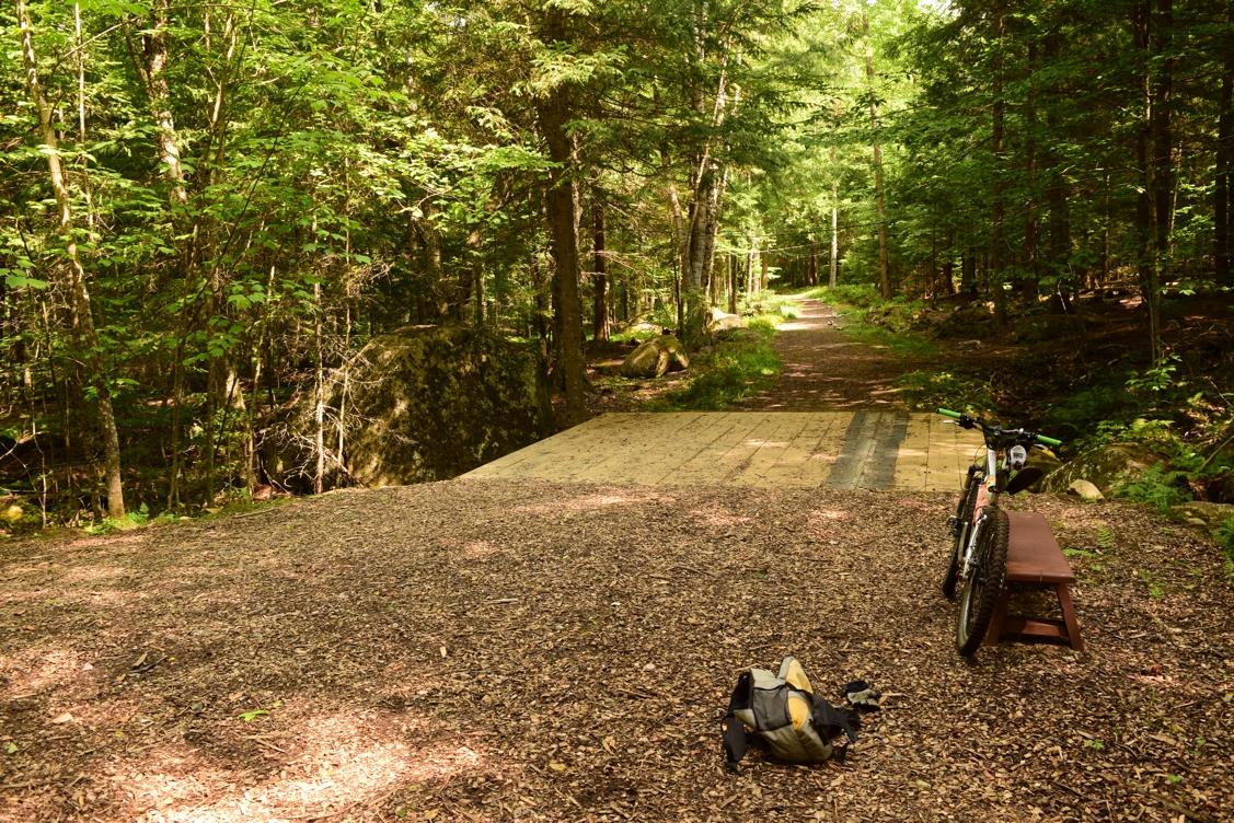 A serene forest pathway with a wooden bridge and a mountain bike parked nearby, alongside a backpack on the ground. Sunlight filters through the lush green trees, illuminating the earthy trail covered in mulch. Dewey Mountain mountain bike trail.