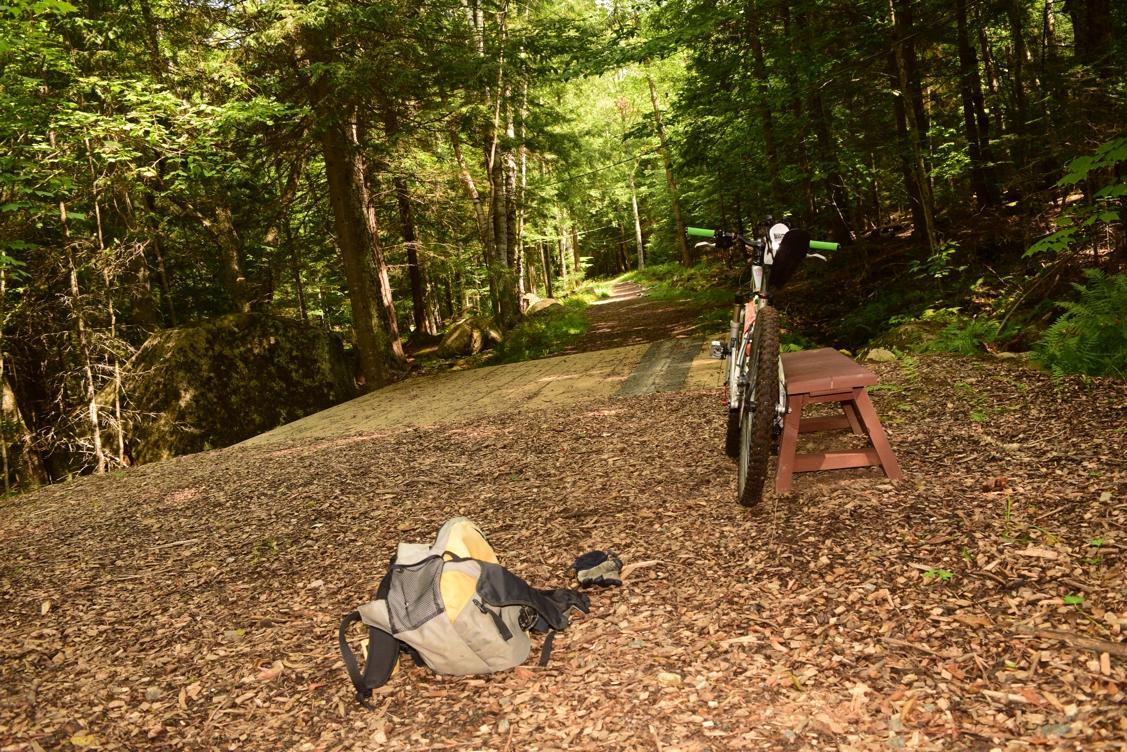 A mountain bike rests on a wooden platform next to a small brown stool, surrounded by a forest of trees and a path leading into the woods. A backpack lies on the ground nearby, with fallen leaves scattered on the forest floor. Dewey Mountain mountain bike trail.