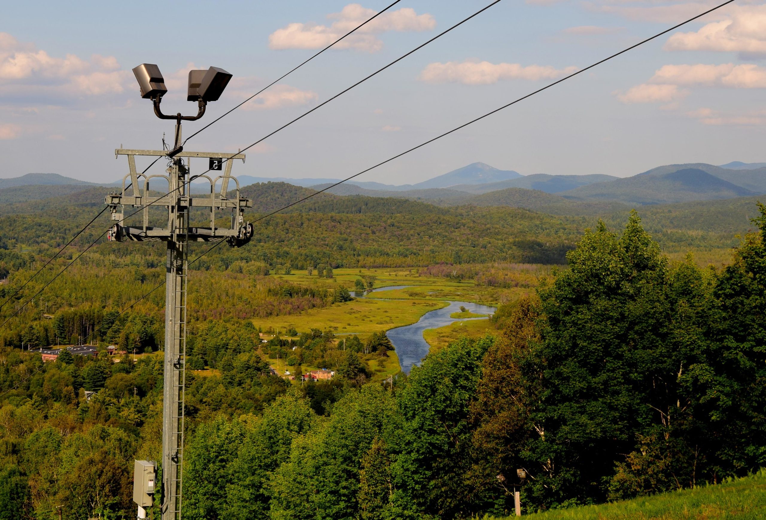 A scenic view from a mountain ridge featuring a chairlift support tower in the foreground. Below, a winding river flows through a lush green valley, surrounded by trees and distant mountains under a partly cloudy sky. Mount Pisgah Ski Area mountain bike trail.