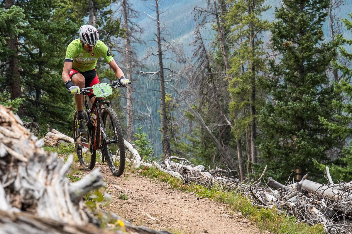 A mountain biker in a green jersey navigates a narrow dirt trail surrounded by trees and fallen branches. The rider is focused, with a determined expression, as they maneuver their bike along the rugged terrain in a mountainous landscape. Colorado Trail: Kenosha Pass To Breckenridge mountain bike trail.