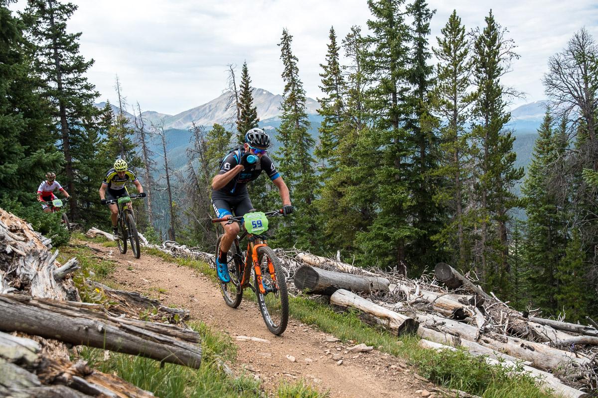 Three mountain bikers navigate a dirt trail surrounded by tall pine trees and scenic mountain views. The riders are wearing helmets and biking gear, with one athlete in the foreground pausing to take a drink. The trail is lined with fallen logs and lush greenery, showcasing an active outdoor adventure in a mountainous landscape. Colorado Trail: Kenosha Pass To Breckenridge mountain bike trail.