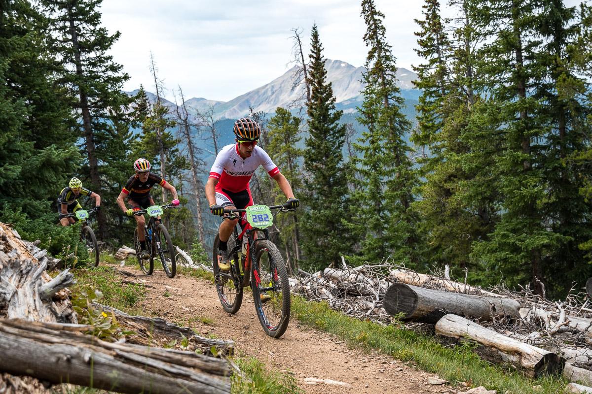 Three mountain bikers navigate a dirt trail surrounded by pine trees and rocky terrain, set against a backdrop of distant mountains. They wear colorful jerseys and helmets while focusing on the path ahead. Colorado Trail: Kenosha Pass To Breckenridge mountain bike trail.