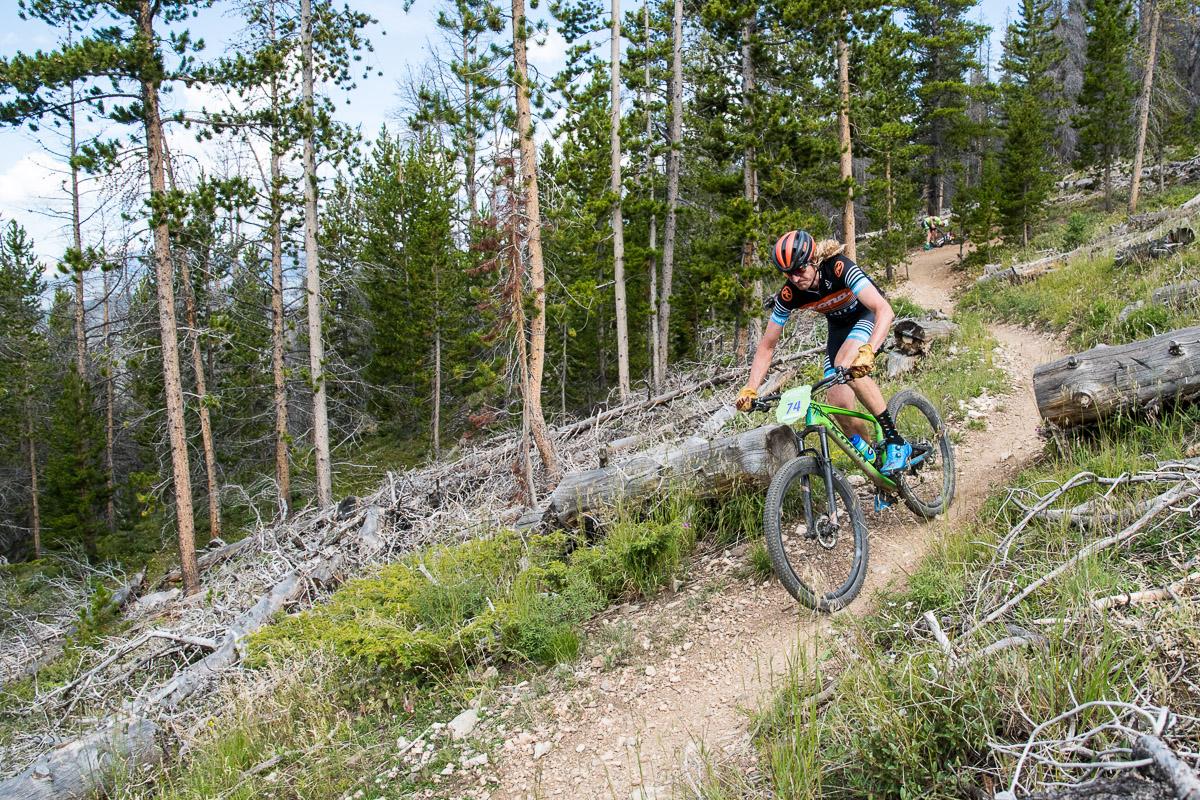 A mountain biker navigates a winding dirt trail amidst a dense forest, maneuvering past fallen logs and greenery on an adventurous ride. Colorado Trail: Kenosha Pass To Breckenridge mountain bike trail.