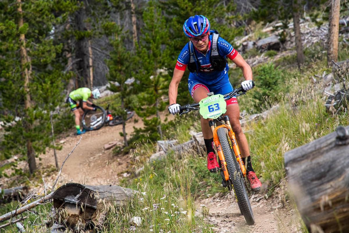 A mountain biker wearing a blue and red racing outfit navigates a dirt trail surrounded by trees, while another cyclist in a green jersey crouches nearby, working on a bicycle. The scene captures a moment during an outdoor cycling event. Colorado Trail: Kenosha Pass To Breckenridge mountain bike trail.