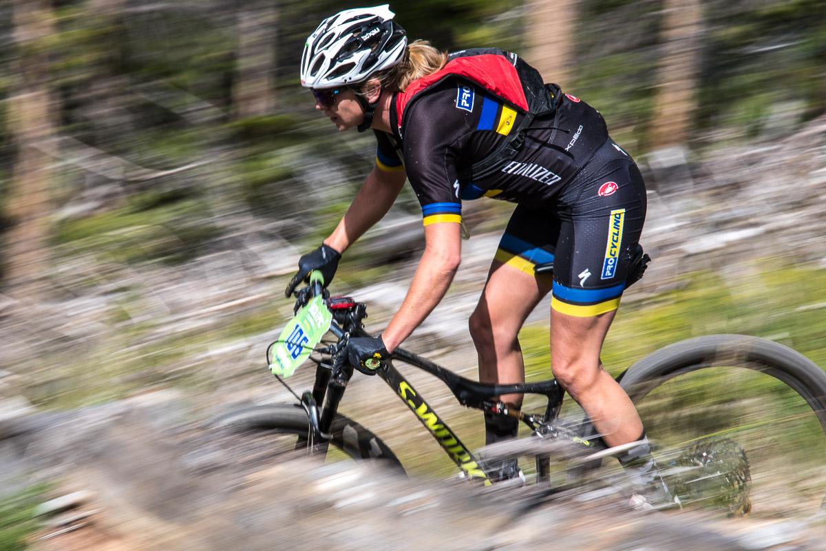 A cyclist in a black and yellow racing outfit rides a mountain bike down a wooded trail, with motion blur indicating high speed. They are wearing a helmet and gloves, and a race number can be seen on the bike. The background features blurred trees and natural landscape, emphasizing the dynamic movement of the scene. Colorado Trail: Kenosha Pass To Breckenridge mountain bike trail.