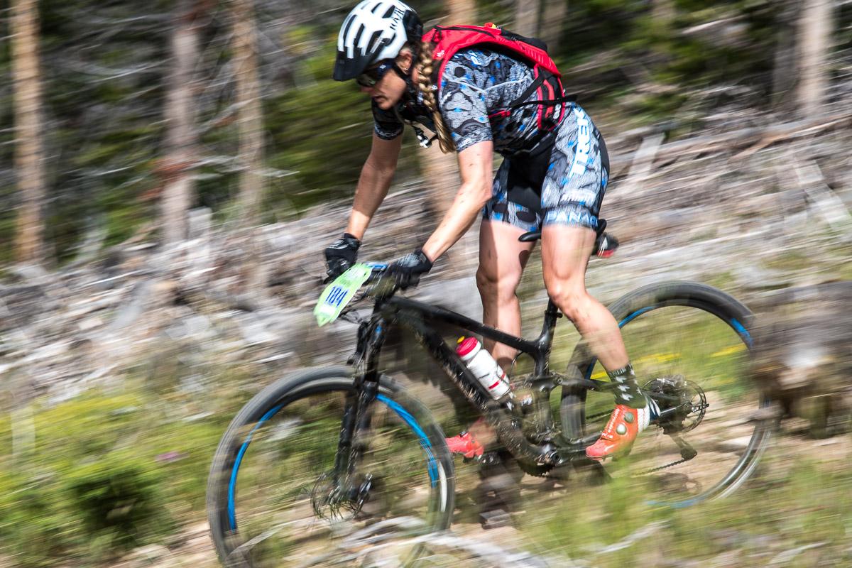 A cyclist in motion navigating a dirt trail, wearing a helmet and a colorful athletic outfit. The bike is equipped for off-road riding, and there is a blurred background of trees and vegetation, indicating speed and movement. The rider has a hydration pack and is focused on the trail ahead. Colorado Trail: Kenosha Pass To Breckenridge mountain bike trail.