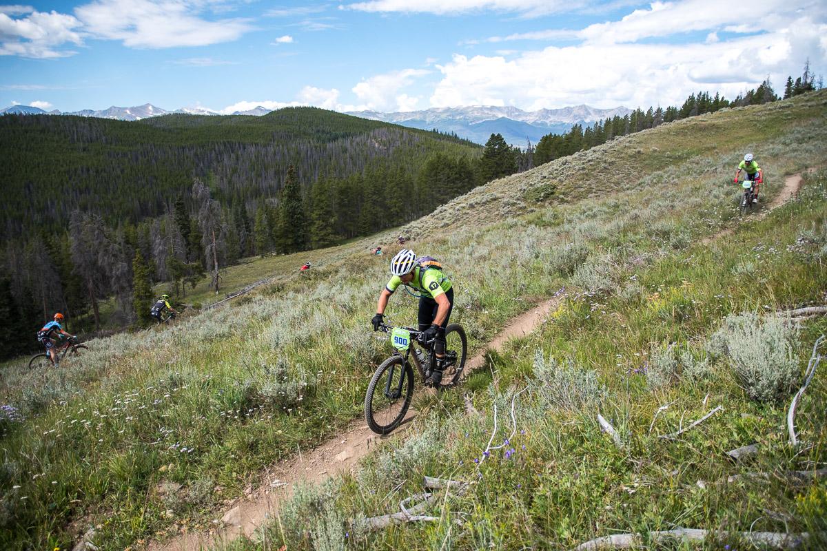 A group of mountain bikers navigating a winding dirt trail through a lush green landscape, surrounded by trees and distant mountains under a partly cloudy sky. The cyclists are wearing bright jerseys and helmets, emphasizing an active outdoor scene. Colorado Trail: Kenosha Pass To Breckenridge mountain bike trail.