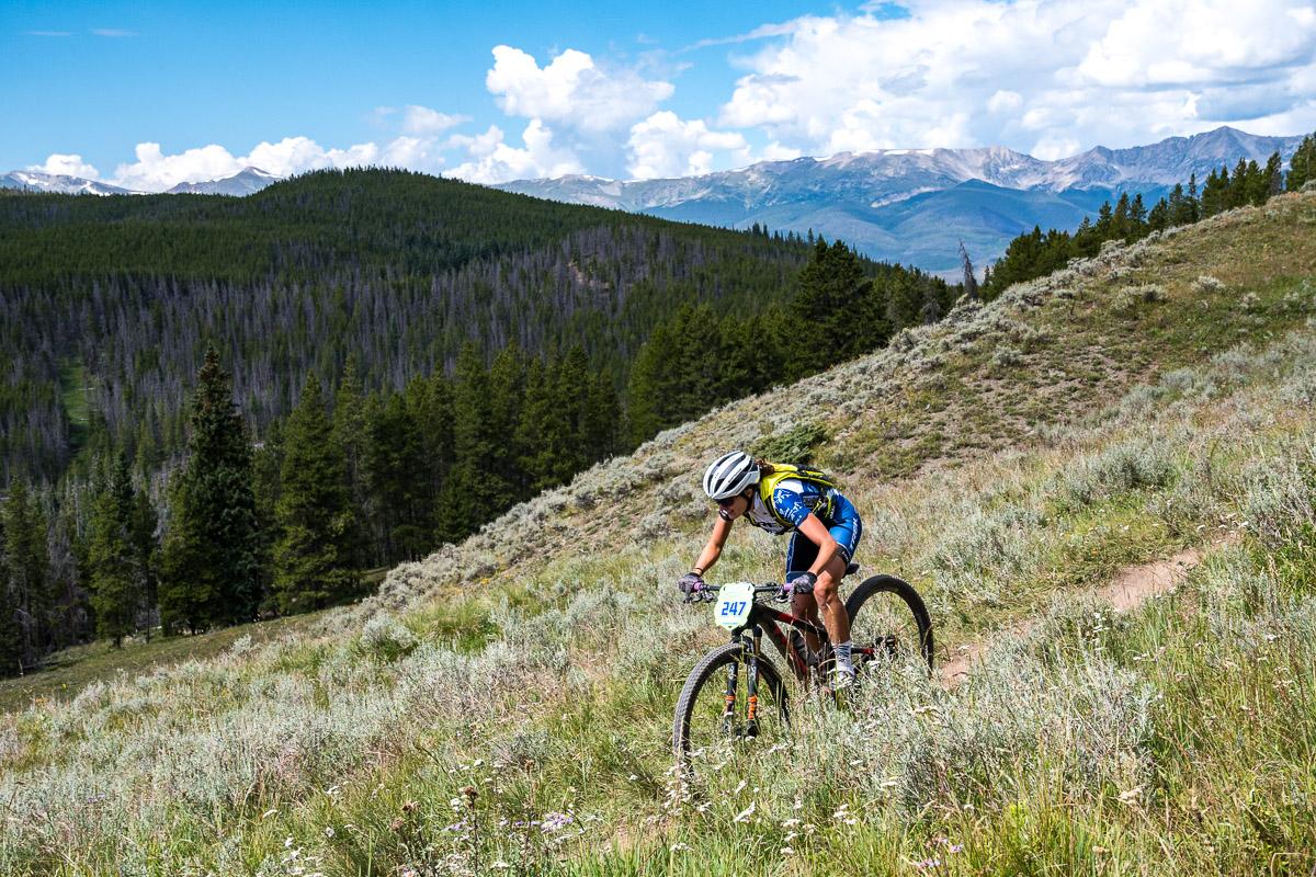 A mountain biker ascends a grassy slope on a trail surrounded by lush trees and mountains under a blue sky with scattered clouds. The cyclist is focused, wearing a helmet and sporting a blue and yellow jersey, with a race number visible on the bike. Colorado Trail: Kenosha Pass To Breckenridge mountain bike trail.