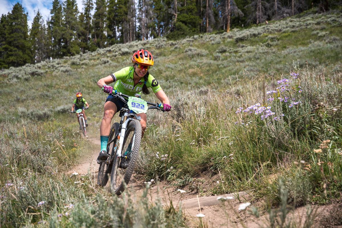 Two women mountain biking on a dirt trail surrounded by green grass and wildflowers. The rider in the foreground, wearing a bright green jersey and helmet, is leaning into a turn, while another rider trails behind in the background. Trees are visible on the hillside in the distance. Colorado Trail: Kenosha Pass To Breckenridge mountain bike trail.