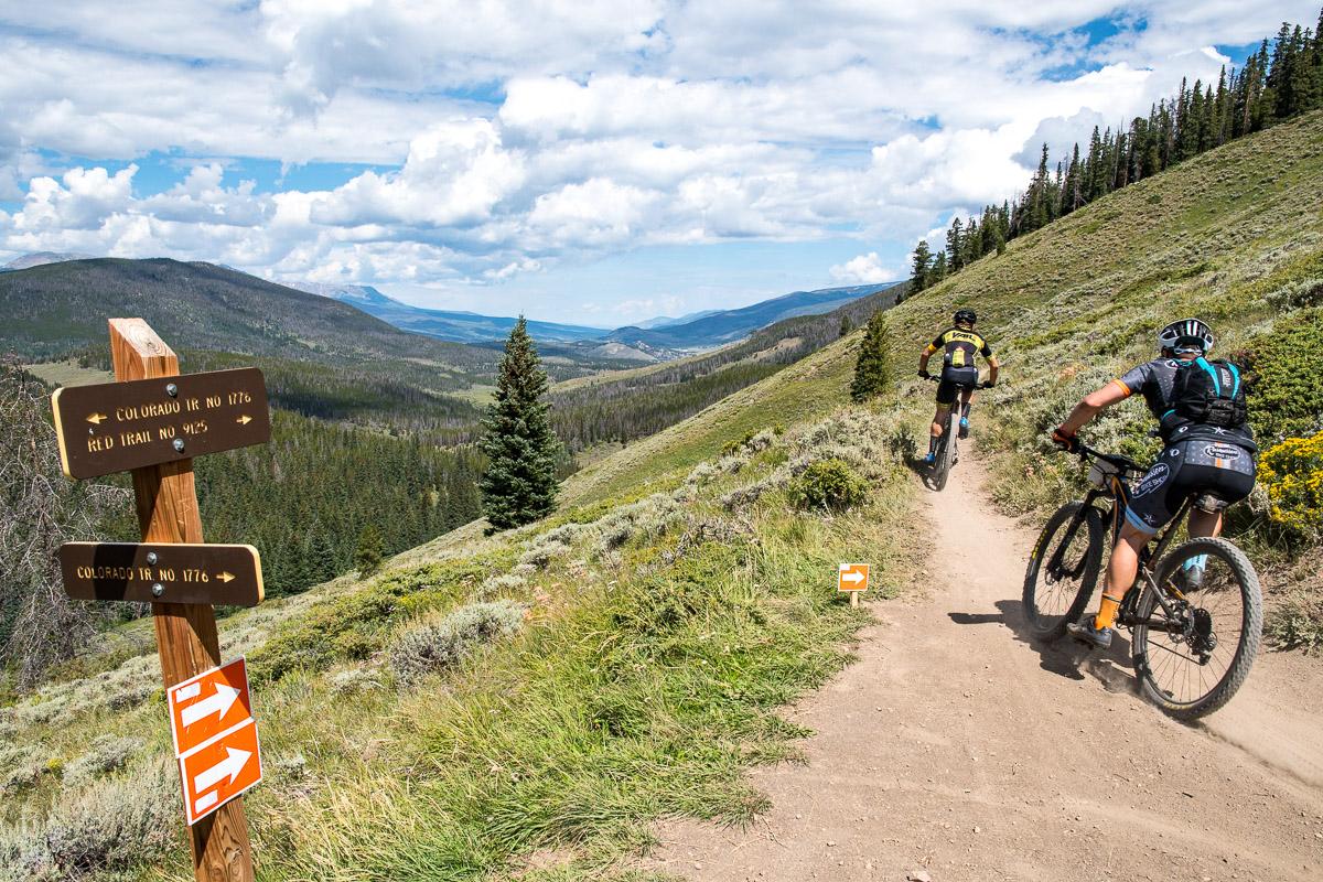 Two mountain bikers riding on a dirt trail through a picturesque mountainous landscape with green hills and trees. There are trail signs indicating different routes. The sky is partly cloudy, and the scene captures a sense of outdoor adventure and exploration. Colorado Trail: Kenosha Pass To Breckenridge mountain bike trail.