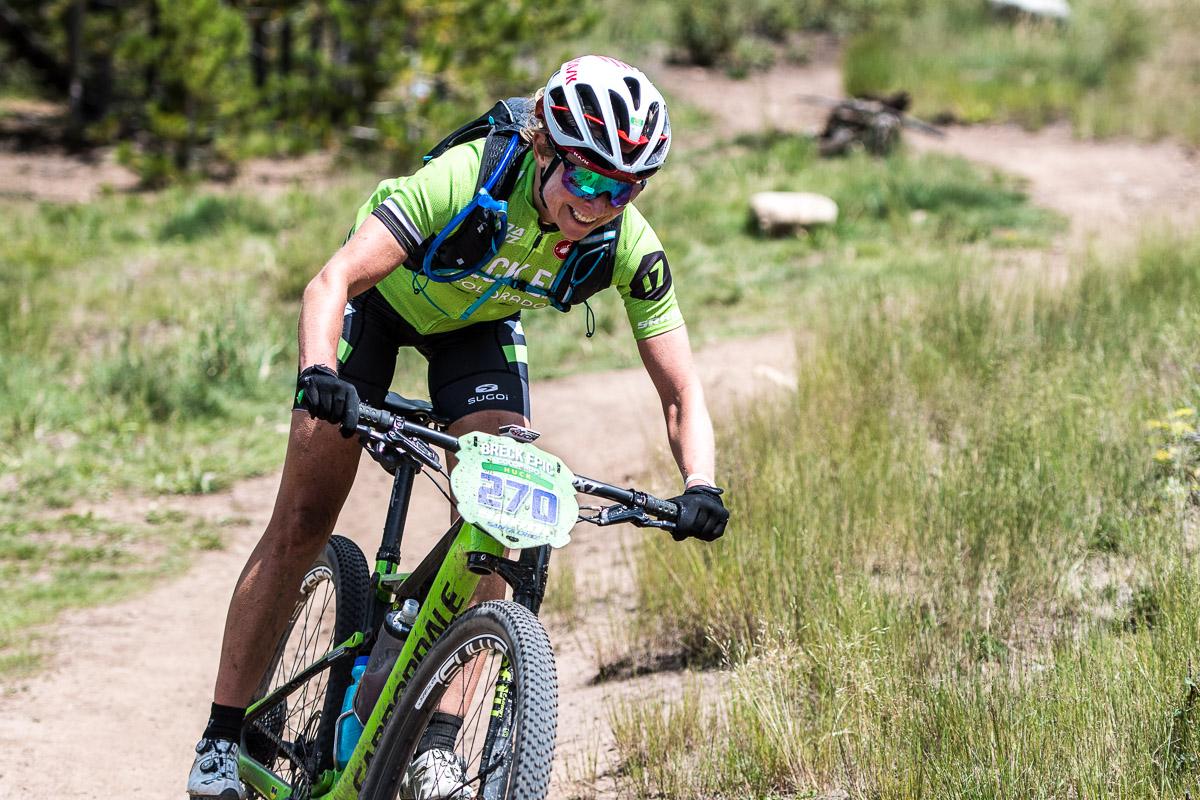 A mountain biker in a bright green and black cycling kit riding on a dirt trail, focused on the path ahead. The rider is wearing a helmet and sunglasses, with a hydration pack on their back, and is climbing uphill through a natural setting with tall grass and trees in the background. A race number plate marked "370" is visible on the bike. Colorado Trail: Kenosha Pass To Breckenridge mountain bike trail.
