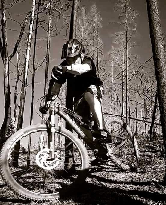A mountain biker riding on a trail among barren, charred trees in a black and white setting. The rider is wearing a helmet and protective gear, leaning forward as they navigate the uneven terrain on a mountain bike. The landscape features tall, skeletal tree trunks, indicating a post-wildfire environment.