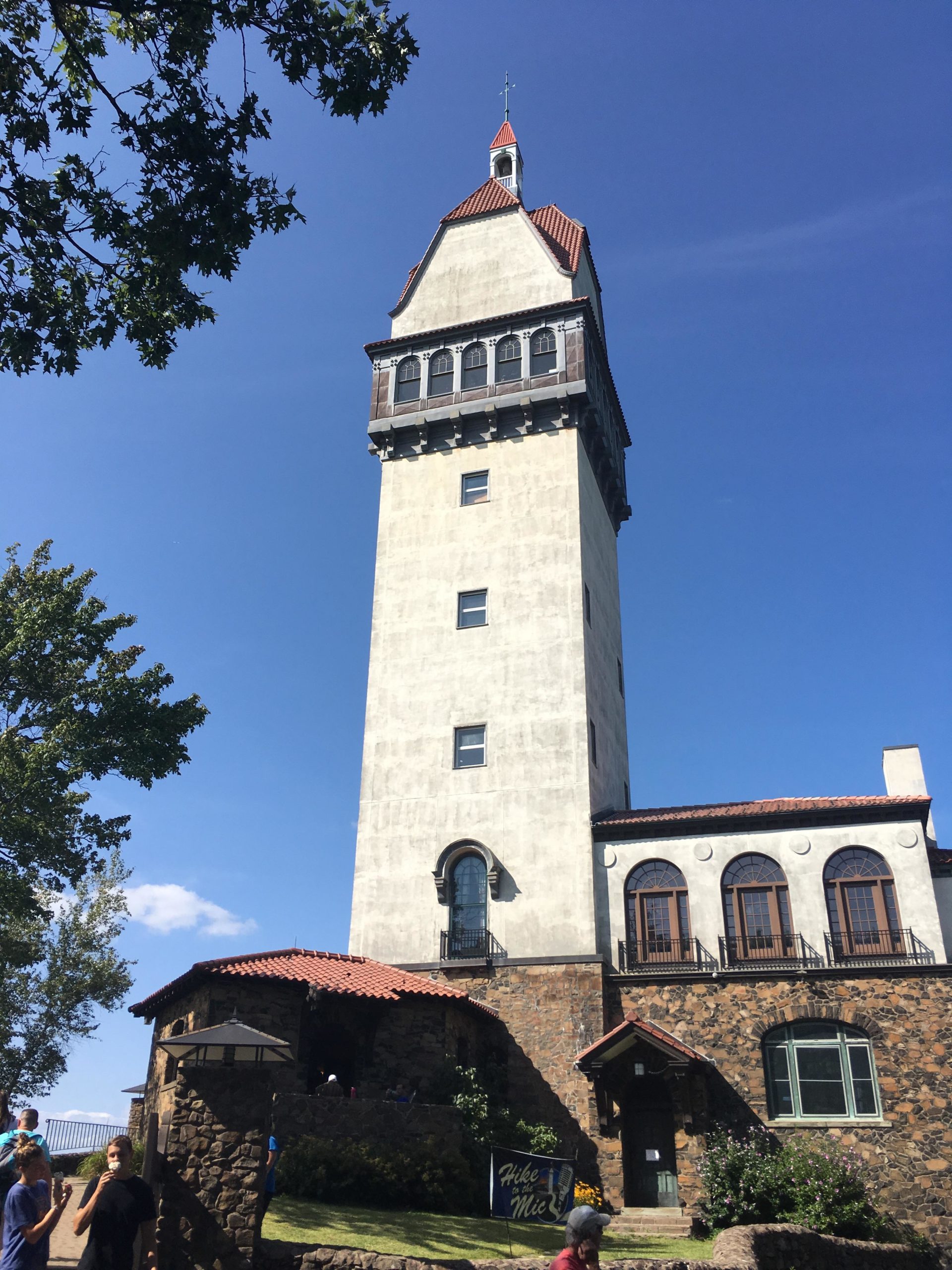 A tall, historic tower with a pointed roof, surrounded by trees and a clear blue sky. The base of the tower features stone architecture, and people can be seen walking nearby. A sign that reads "Hike" is visible in the foreground, indicating a scenic area for outdoor activities. Heublein Tower mountain bike trail.