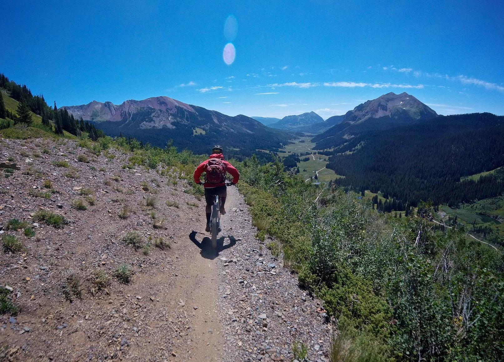 A mountain biker rides along a rocky trail with stunning views of rolling hills and mountain ranges under a clear blue sky. Lush green trees line the path, and sunlight illuminates the landscape. Trail 401 mountain bike trail.