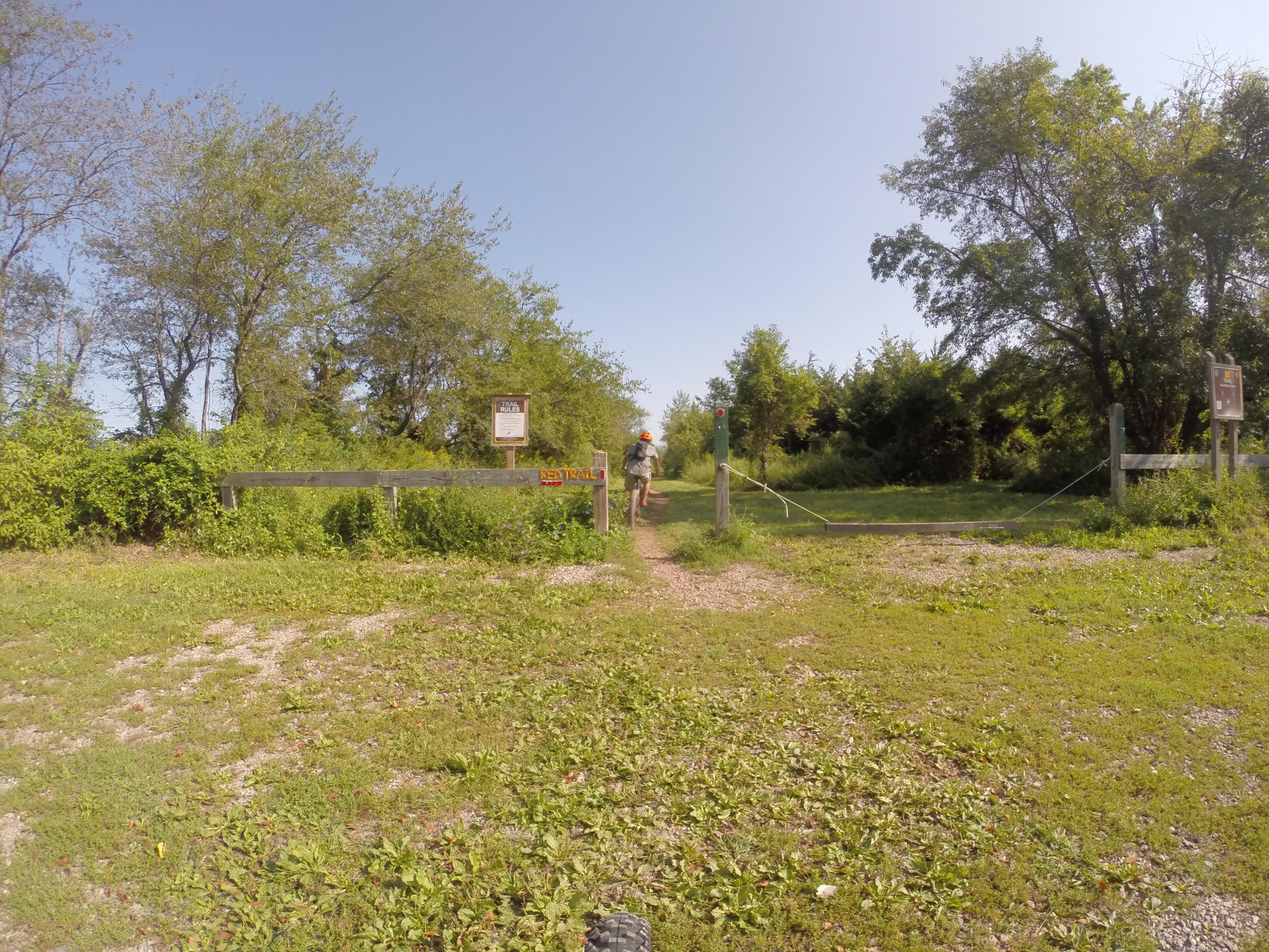 A person walking along a grassy trail leading into a wooded area, with a sign indicating the entrance to a trail. The landscape features trees and shrubs in the background under a clear blue sky. Six Mile Run mountain bike trail.
