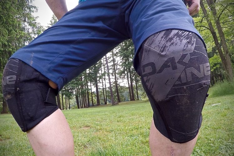 Close-up view of a person's legs wearing protective knee pads in an outdoor setting, surrounded by grass and trees. The knee pads are black with a logo, showing signs of use. The person is wearing blue shorts and is positioned in a way that highlights the knee protection gear.